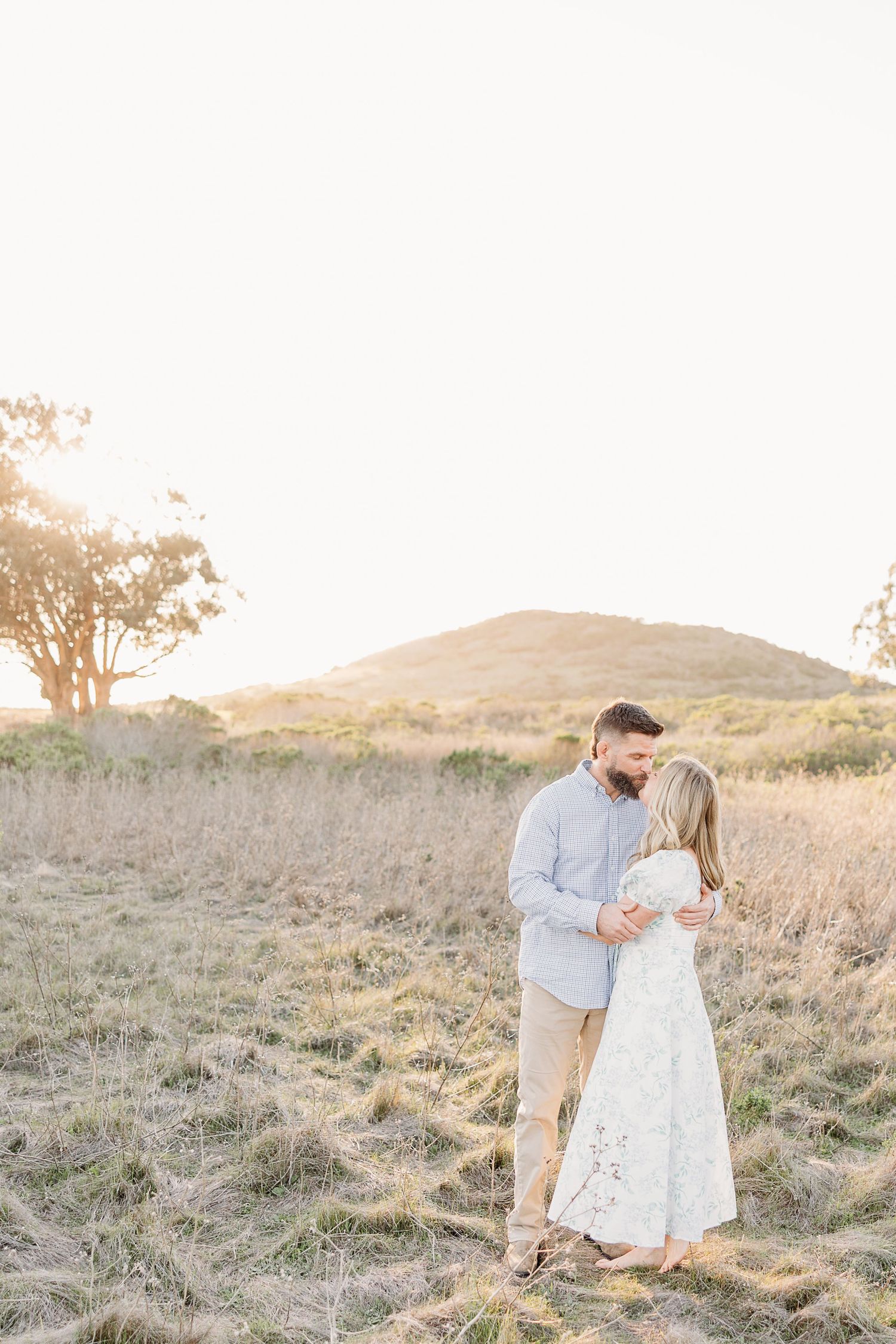 A couple in white and gray attire share a romantic moment in a sunlit grassy field at sunset.
