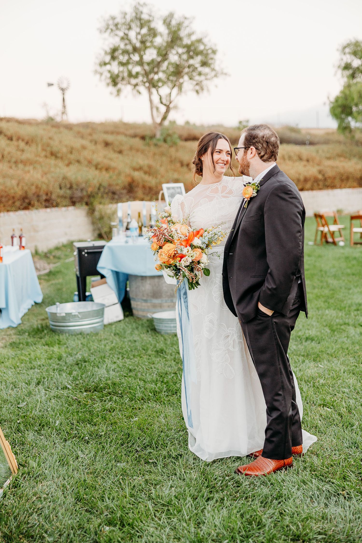 An outdoor wedding celebration featuring a beautiful white dress and dark suit with cheerful orange accents on a grassy lawn.