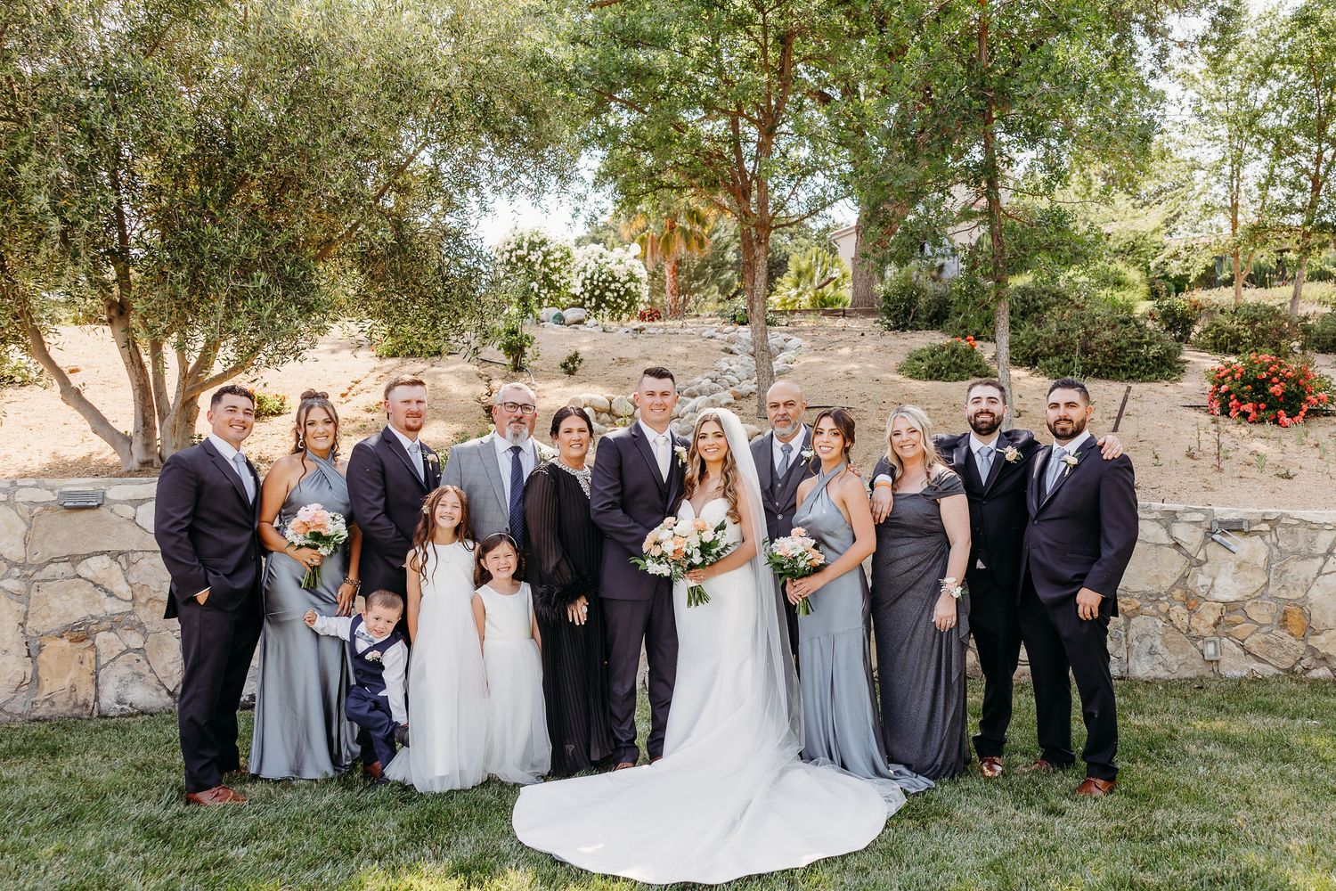 Wedding party in coordinated gray and navy attire poses together on landscaped lawn with stone wall backdrop.