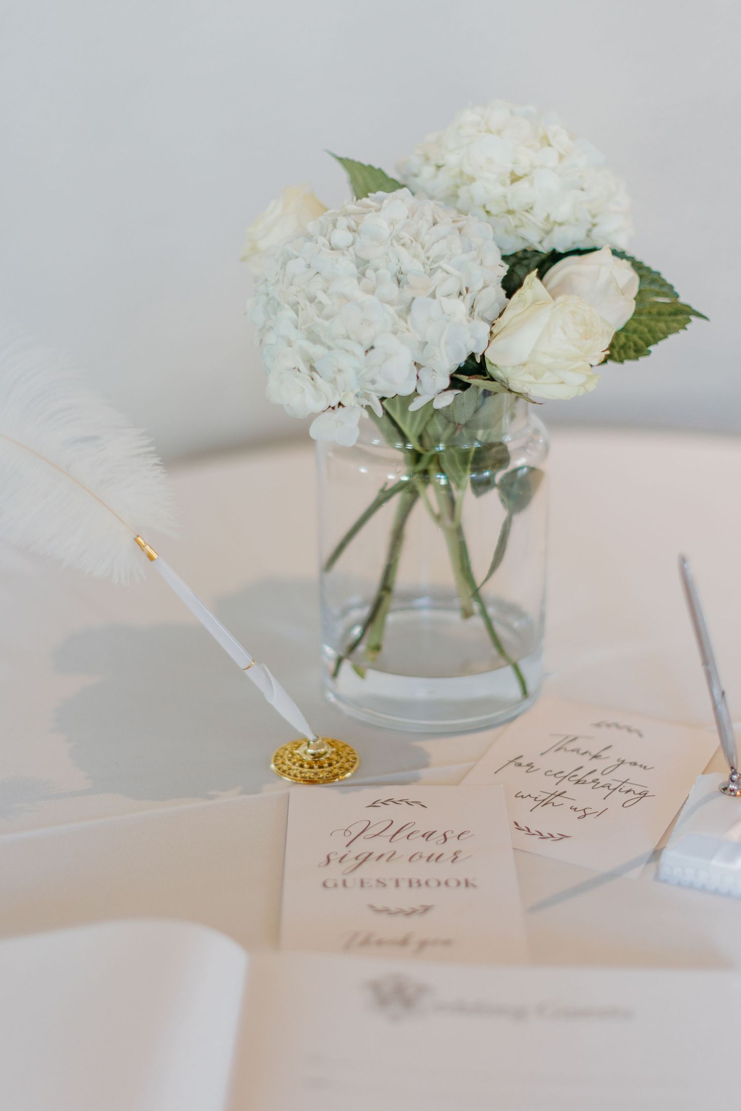 White hydrangeas and roses in a glass vase next to a feather pen and wedding invitation.