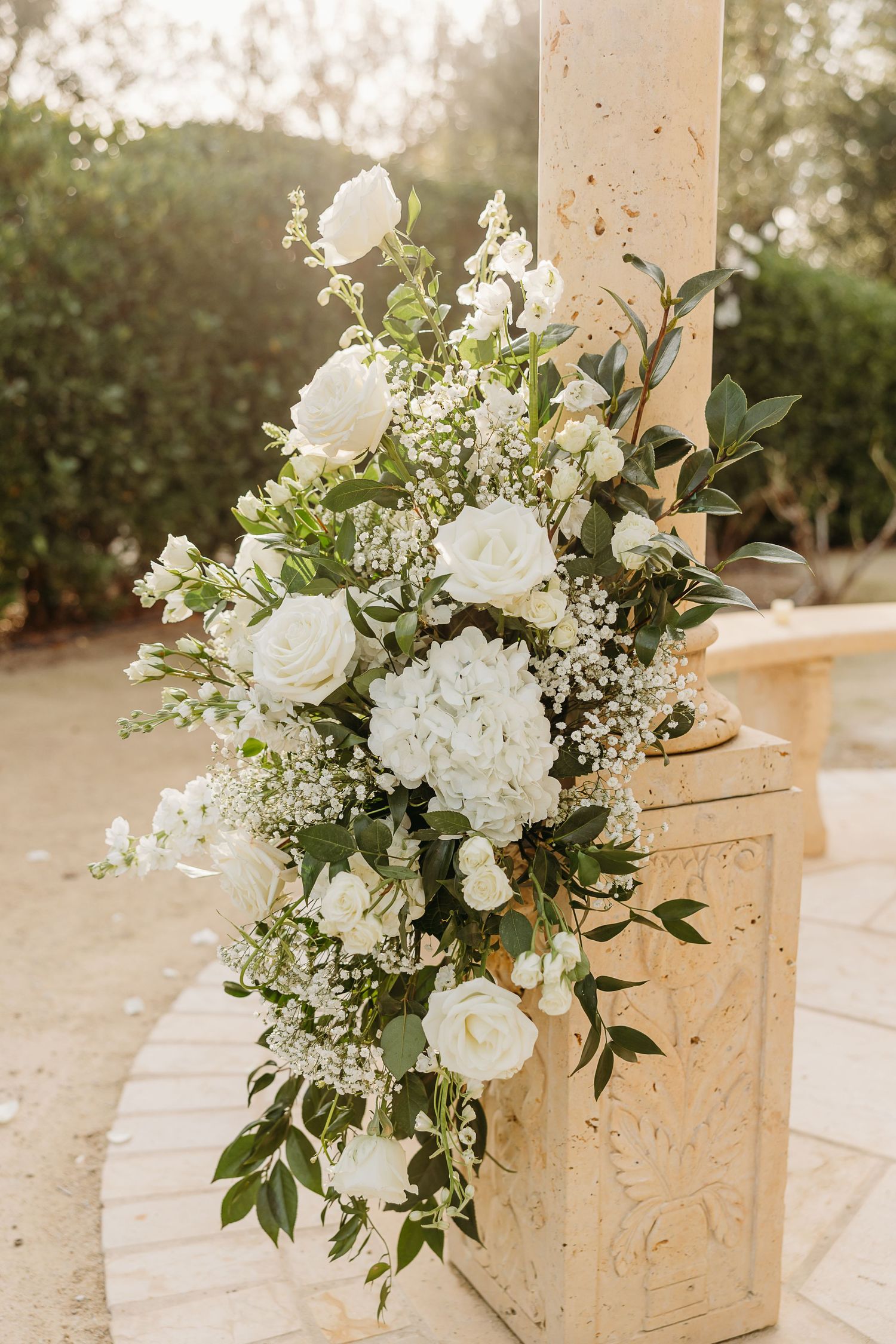 Elegant white floral arrangements with roses and greenery decorate wooden boxes at an outdoor wedding venue.