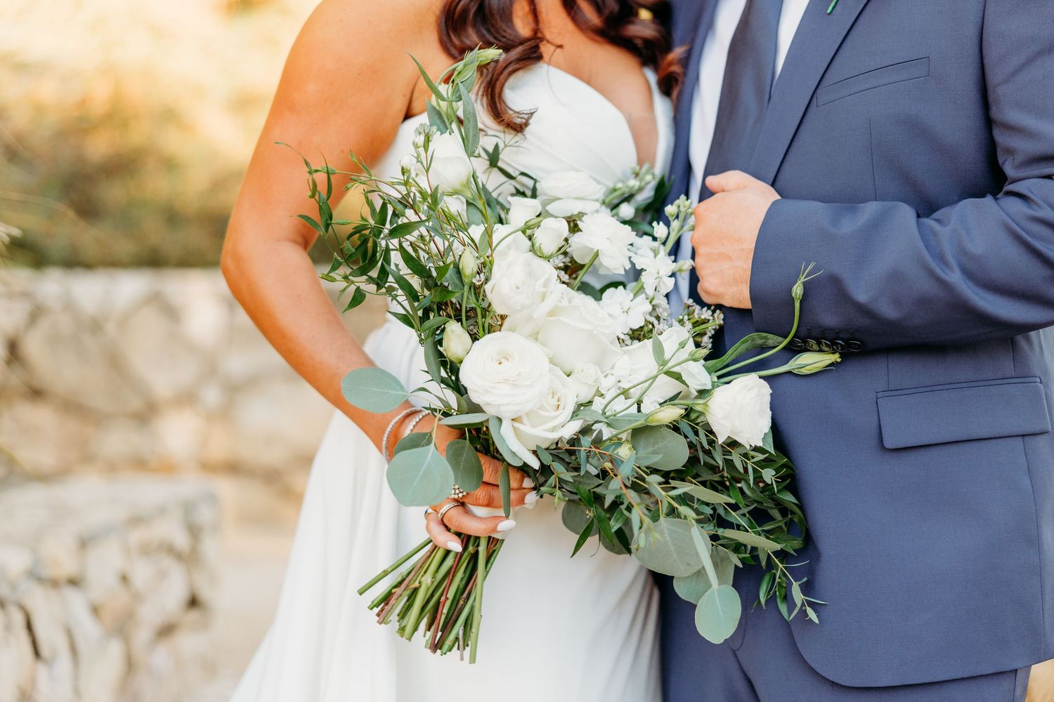 Close-up of wedding couple holding elegant white and green floral bouquet with eucalyptus against stone wall.