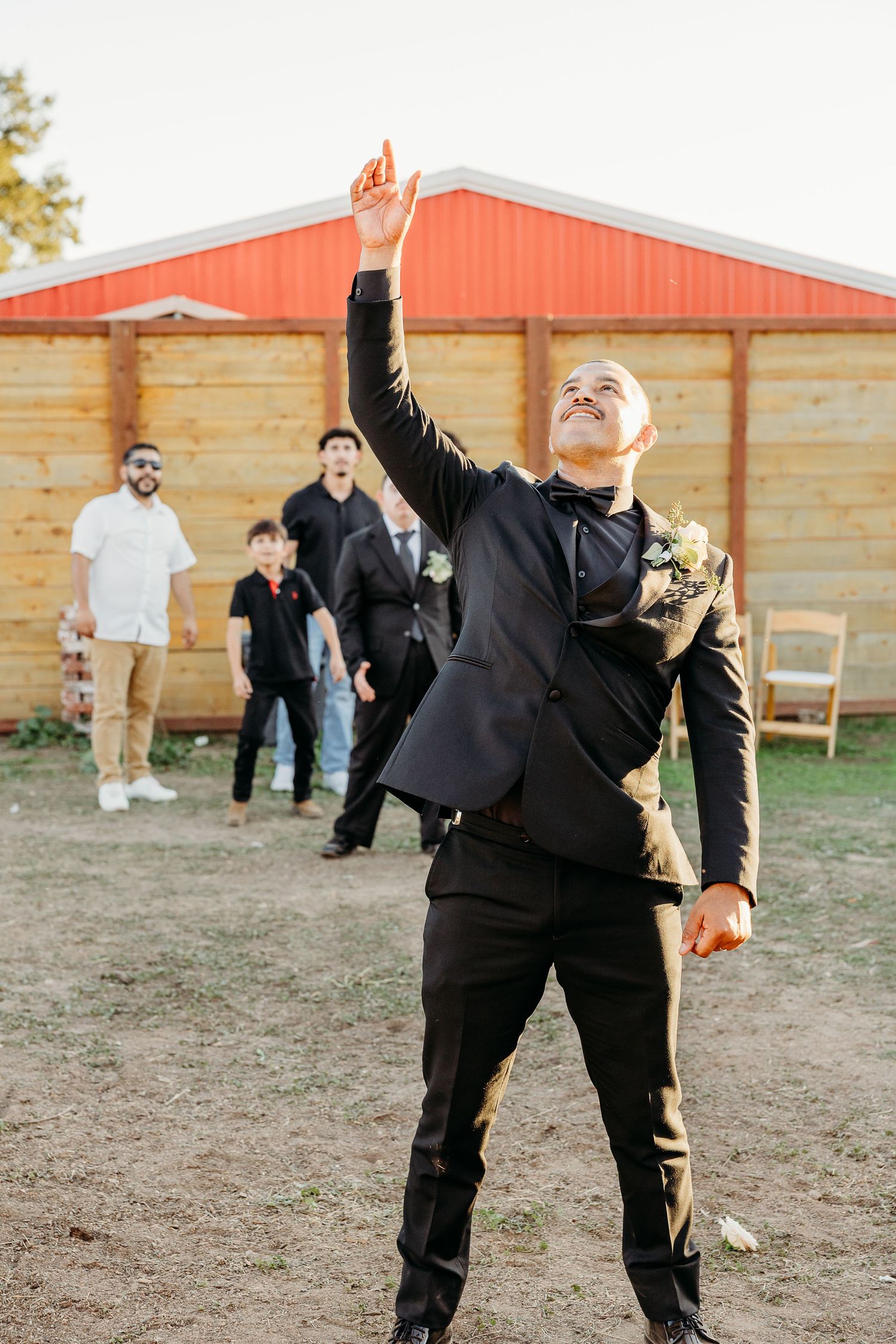 Groomsmen in black suits playing football outside a red barn during wedding festivities.