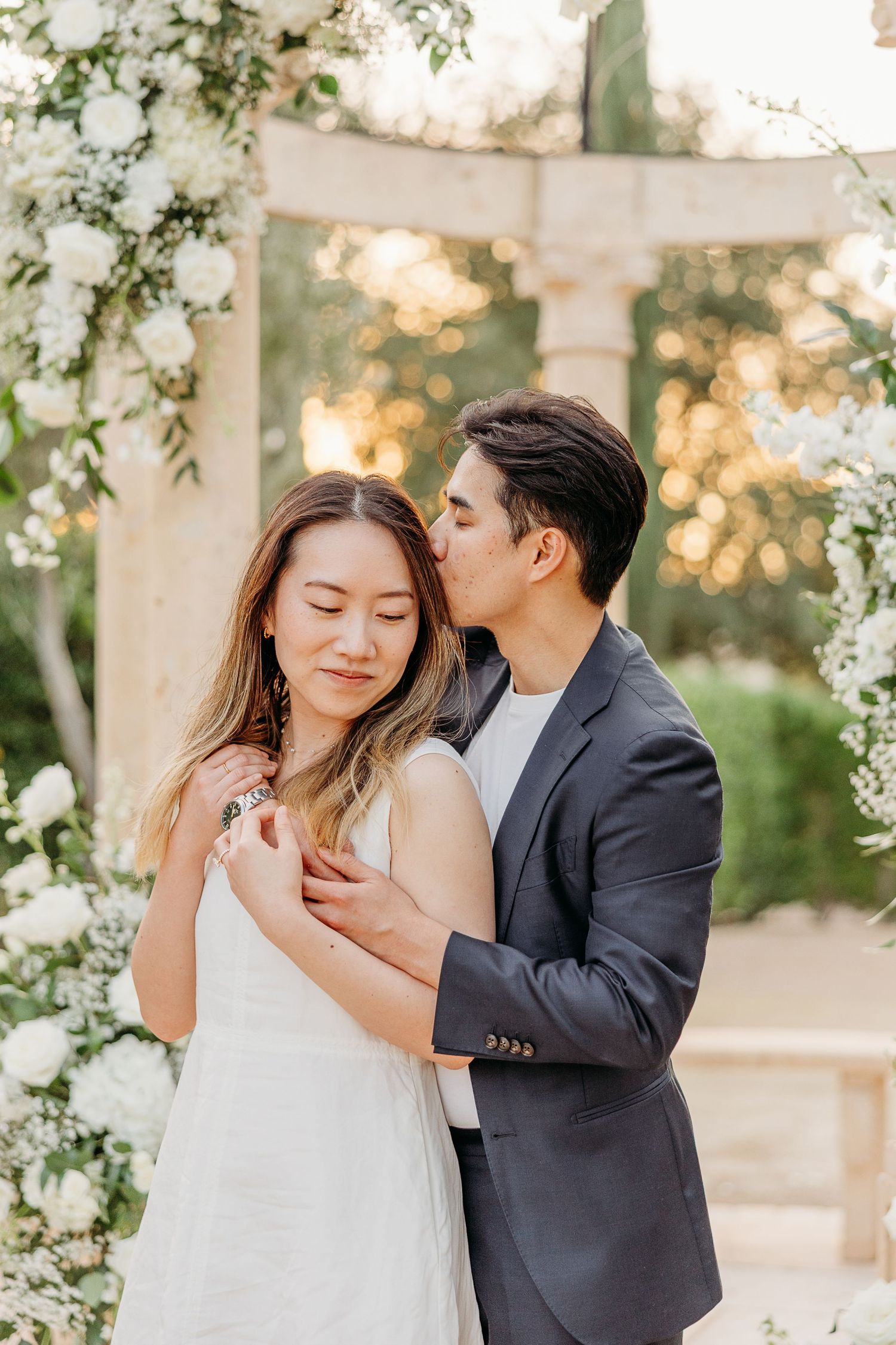 Tender moment captured under white floral arch during romantic photoshoot.