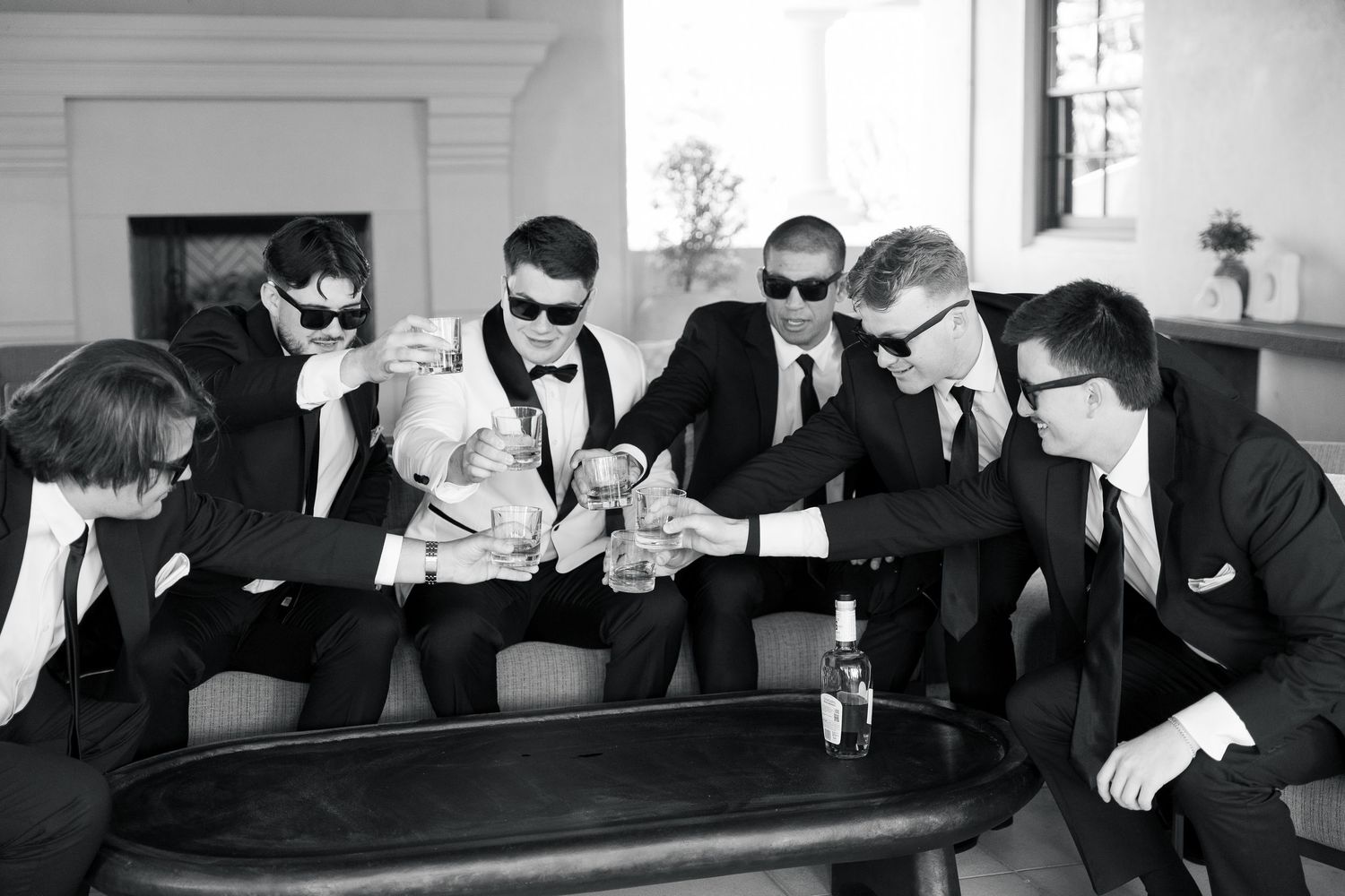 Groomsmen in black suits and sunglasses toasting with drinks at a wedding celebration.