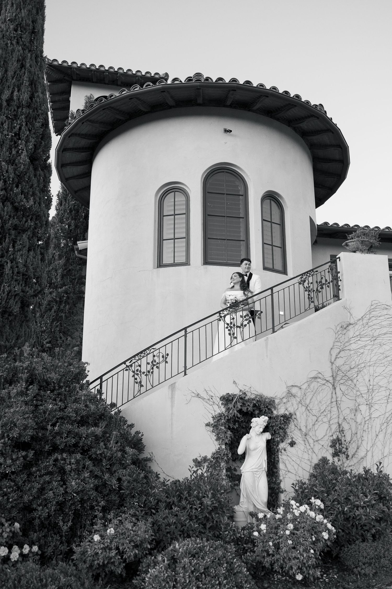 Bride and groom pose on balcony of elegant Spanish-style villa in black and white photo.