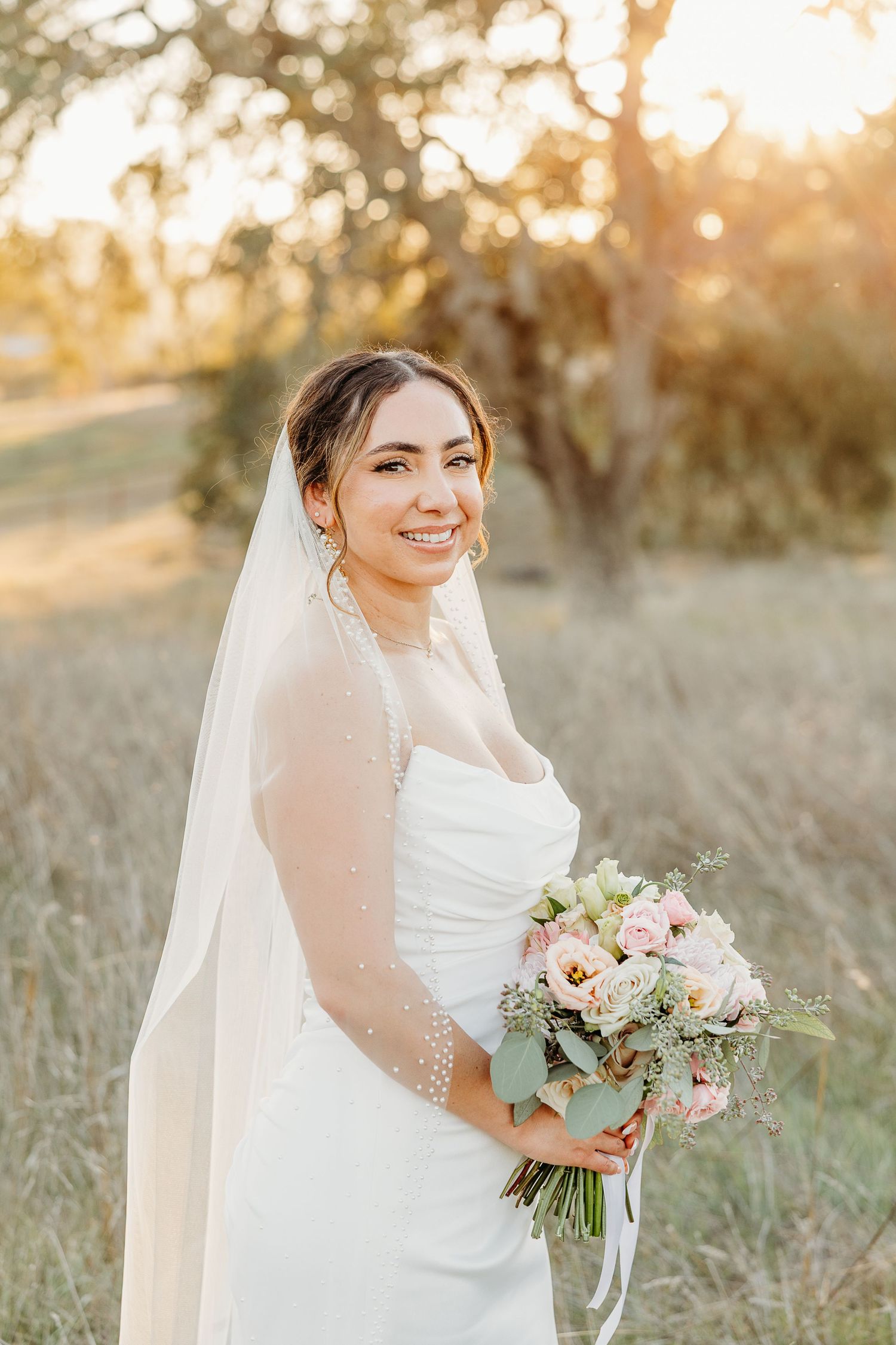 Bridal portraits in a golden field at sunset with a bouquet of pink and white flowers.
