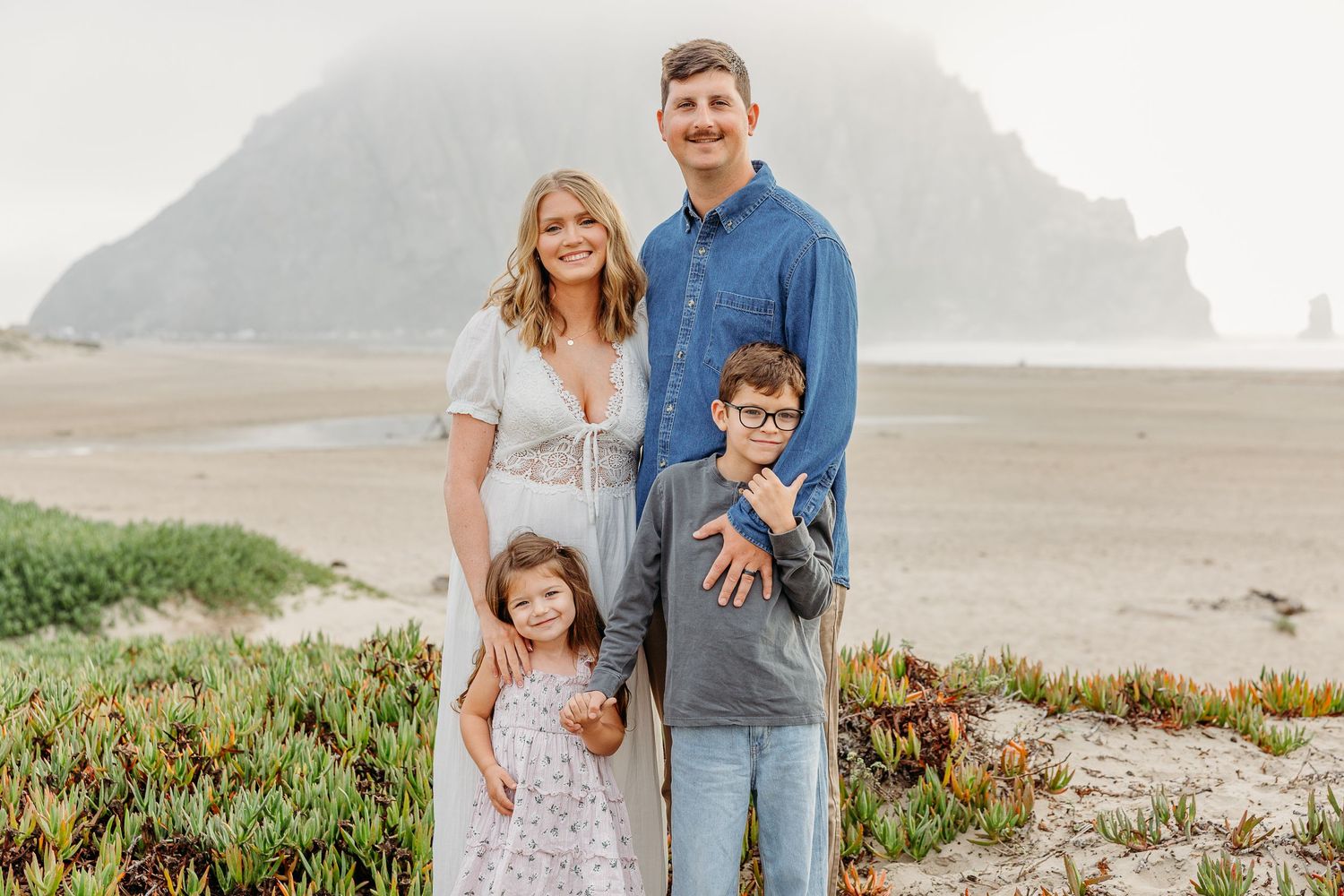 Family photoshoot at a scenic coastal location with mountains and beach vegetation.