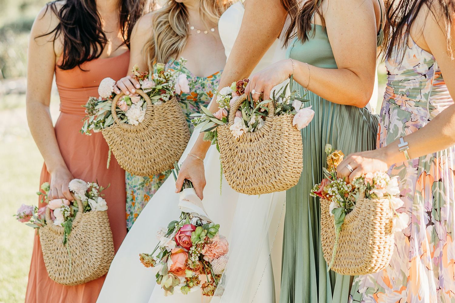 Bridesmaids in pastel dresses hold woven straw baskets filled with pink and peach floral arrangements.