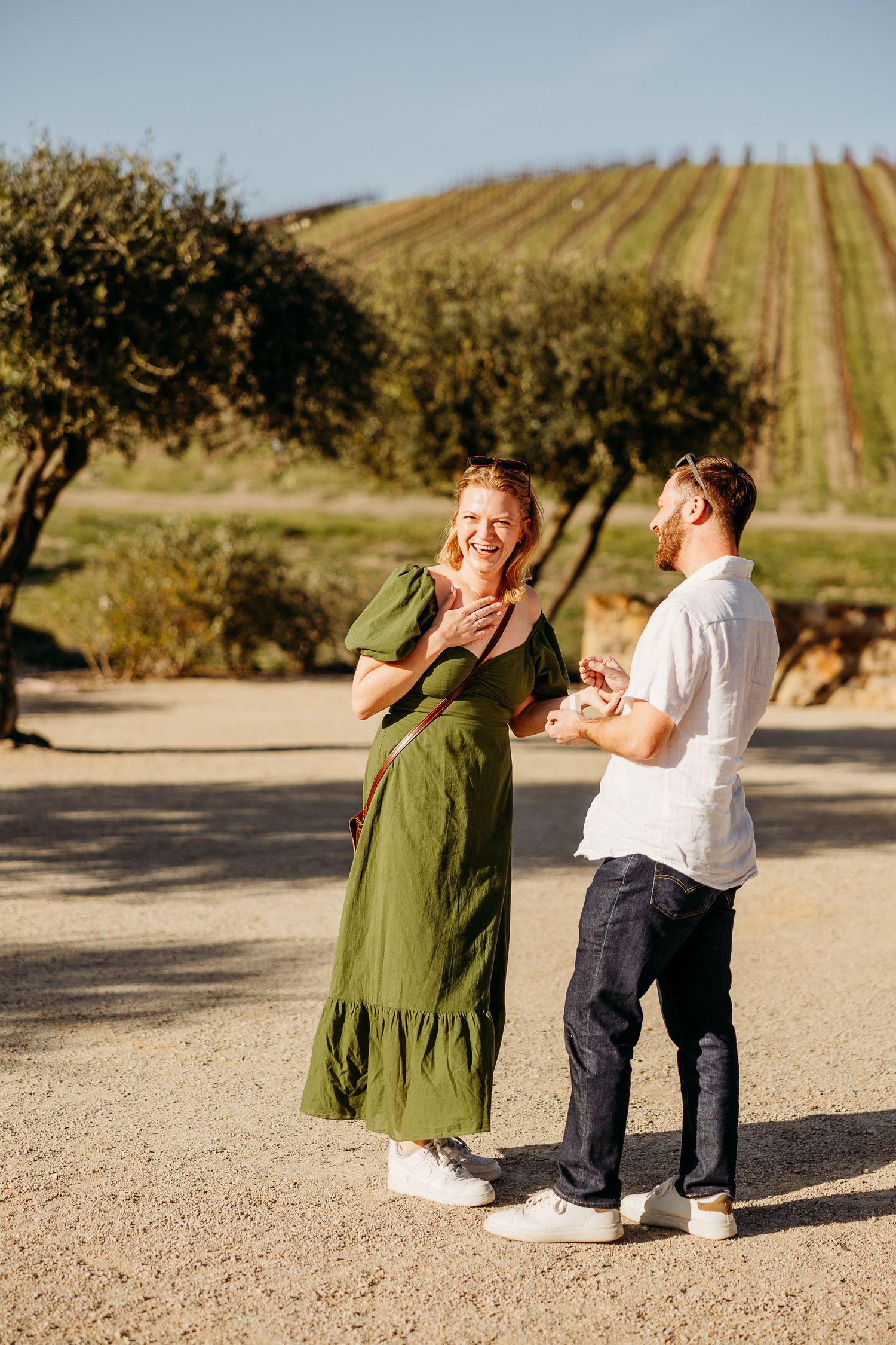 A couple shares a lighthearted moment on a dirt path with rolling hills in the background.