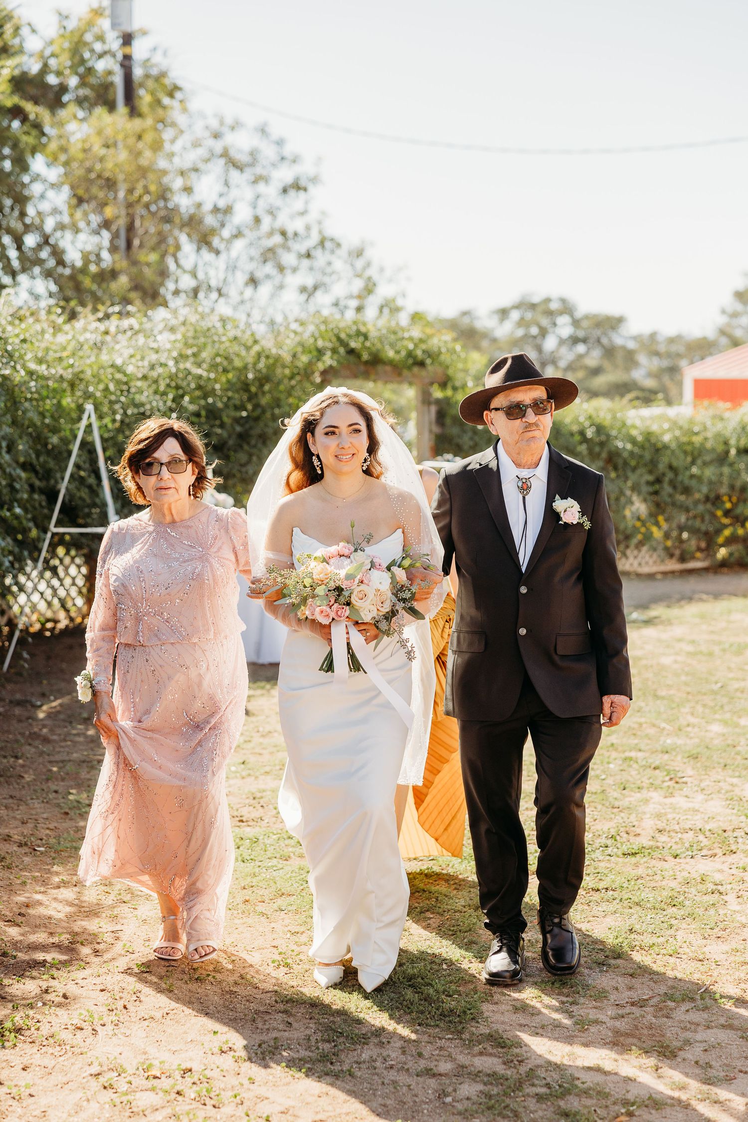 Wedding party walking along a sunlit outdoor path with the bride in white and bridesmaids in pink.
