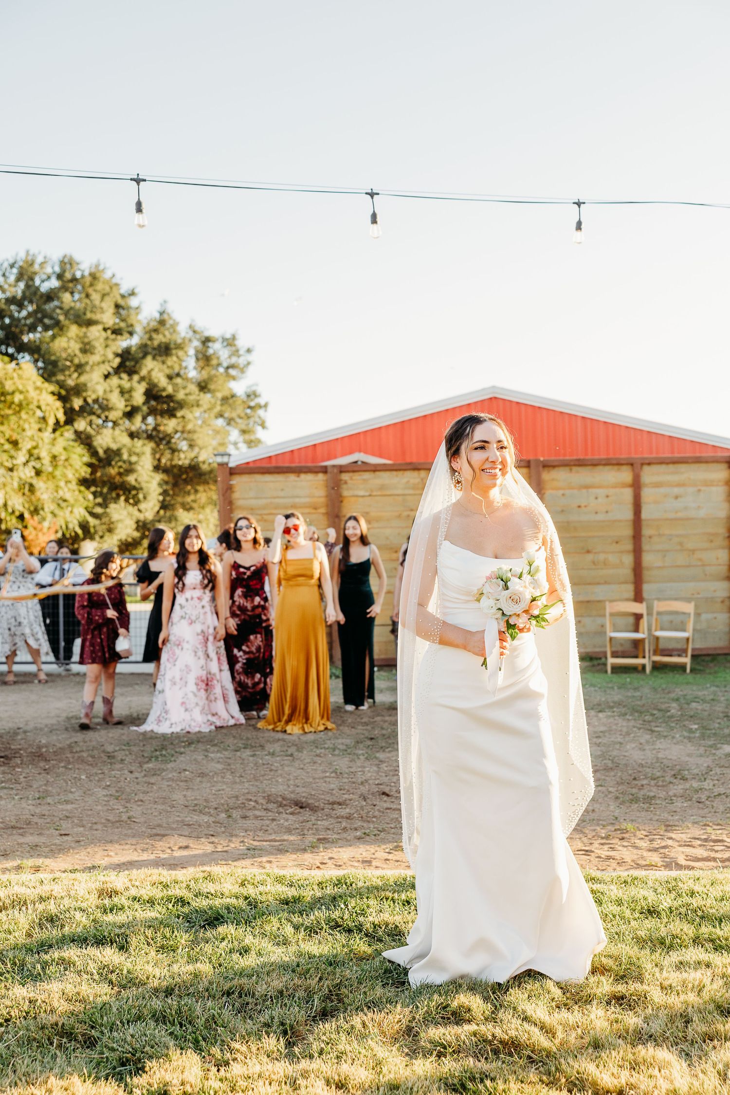 Wedding party stands on grassy field at sunset while bride in white dress poses in golden hour light.