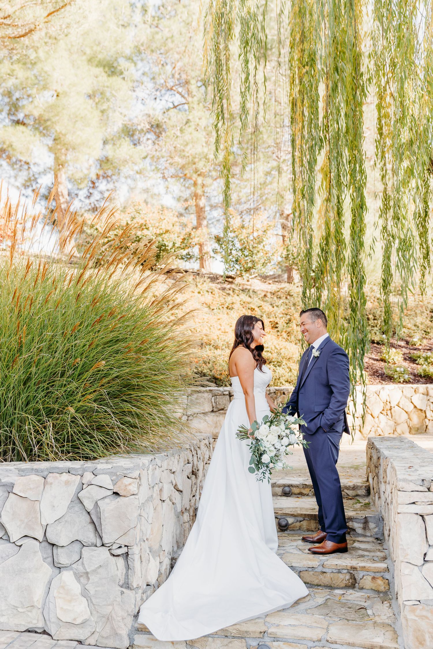 Wedding couple poses against stone wall with willow trees in romantic outdoor setting.