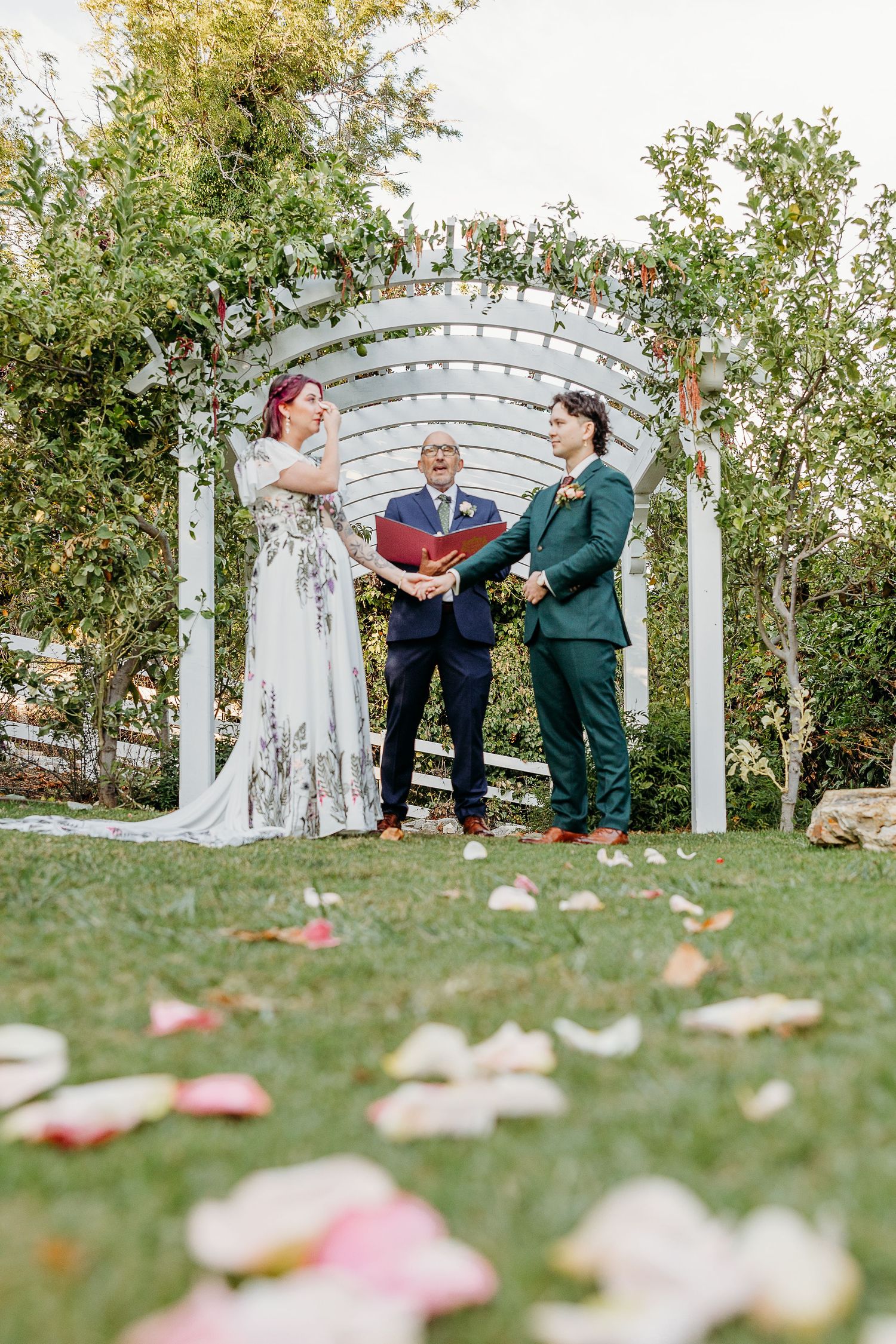 Wedding ceremony setup outdoors with white arbor and scattered rose petals on grass.