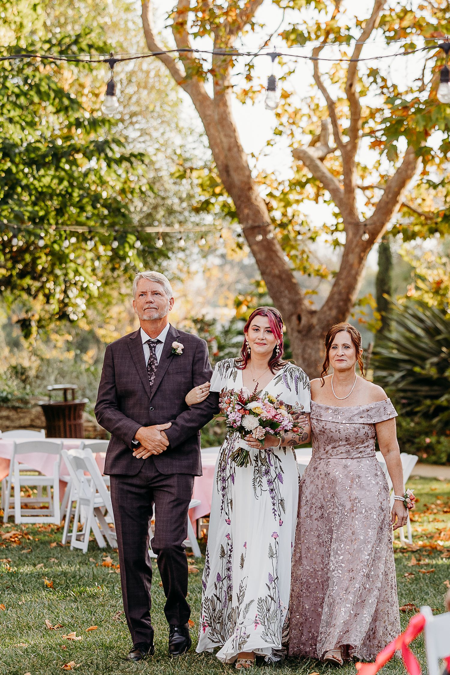 Wedding party walking down garden path with autumn trees and white chairs in background.