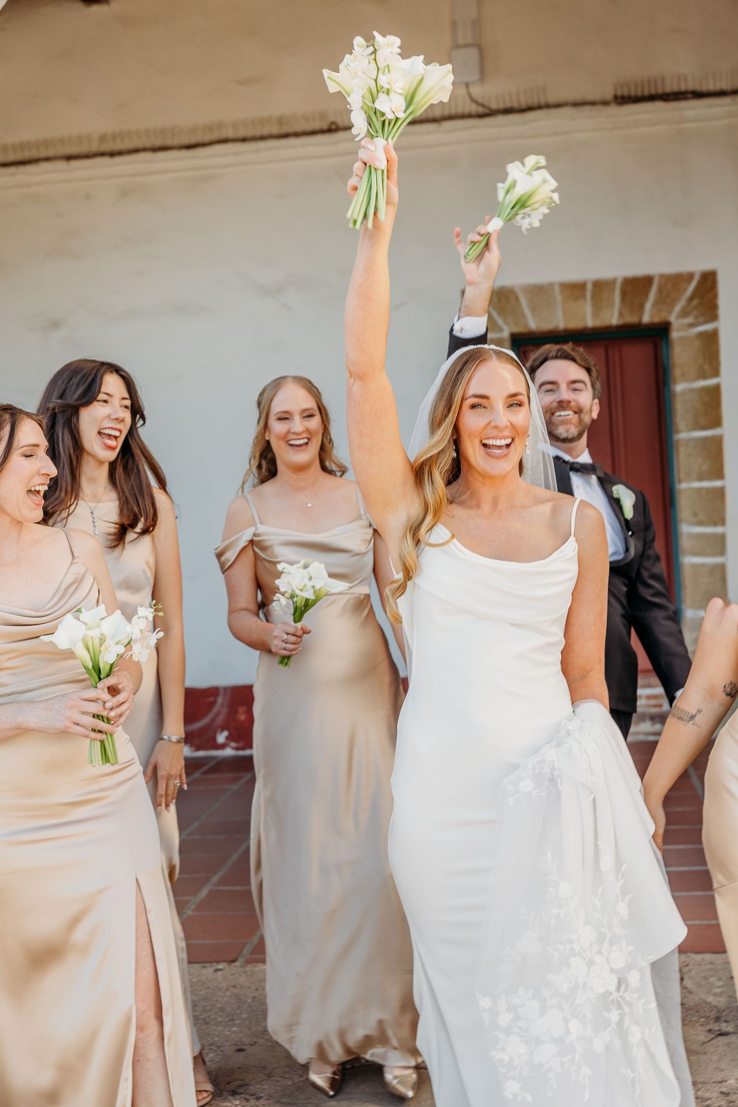 Joyful wedding party celebrates with white floral bouquets against neutral wall backdrop.