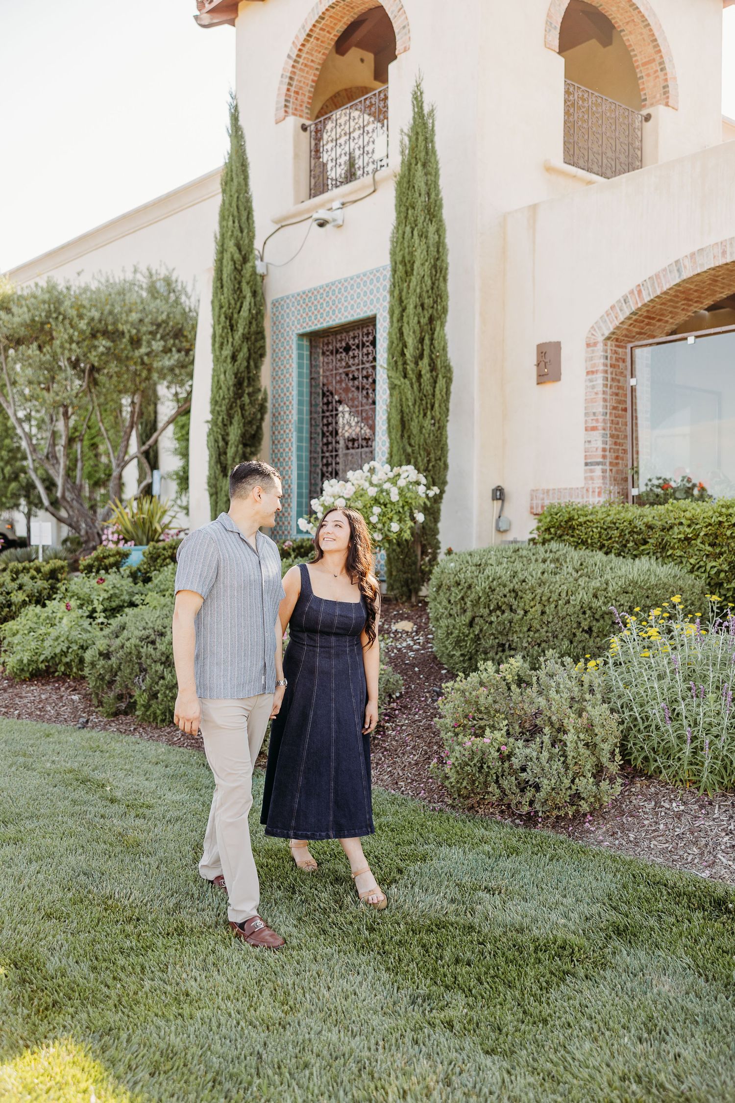 A couple takes a romantic stroll along a Mediterranean-style building with cypress trees and manicured gardens.