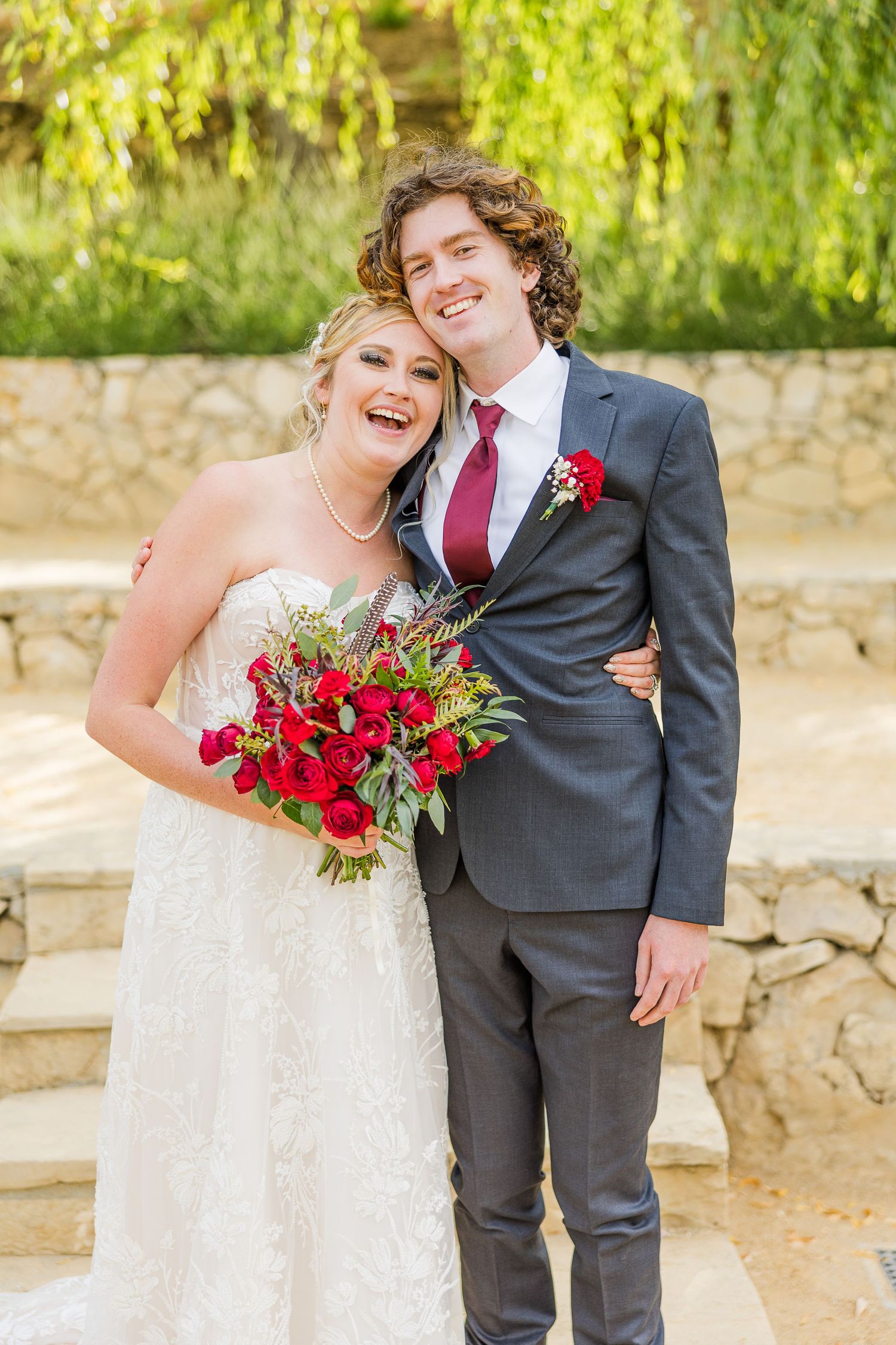 Joyful couple shares romantic moment in formal wedding attire with red floral accents.