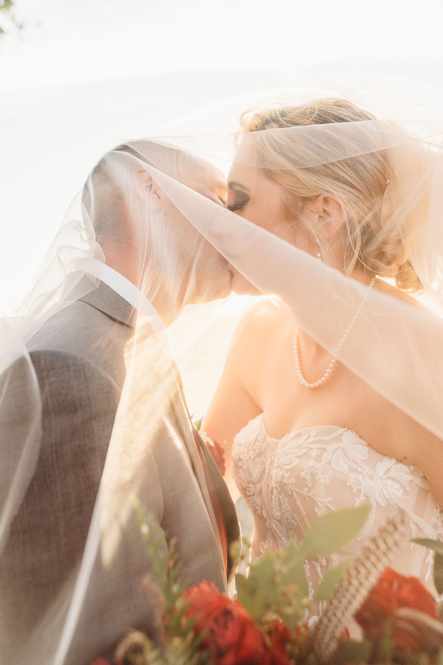 A romantic wedding portrait with a couple sharing a tender kiss under a delicate veil in warm sunlight.
