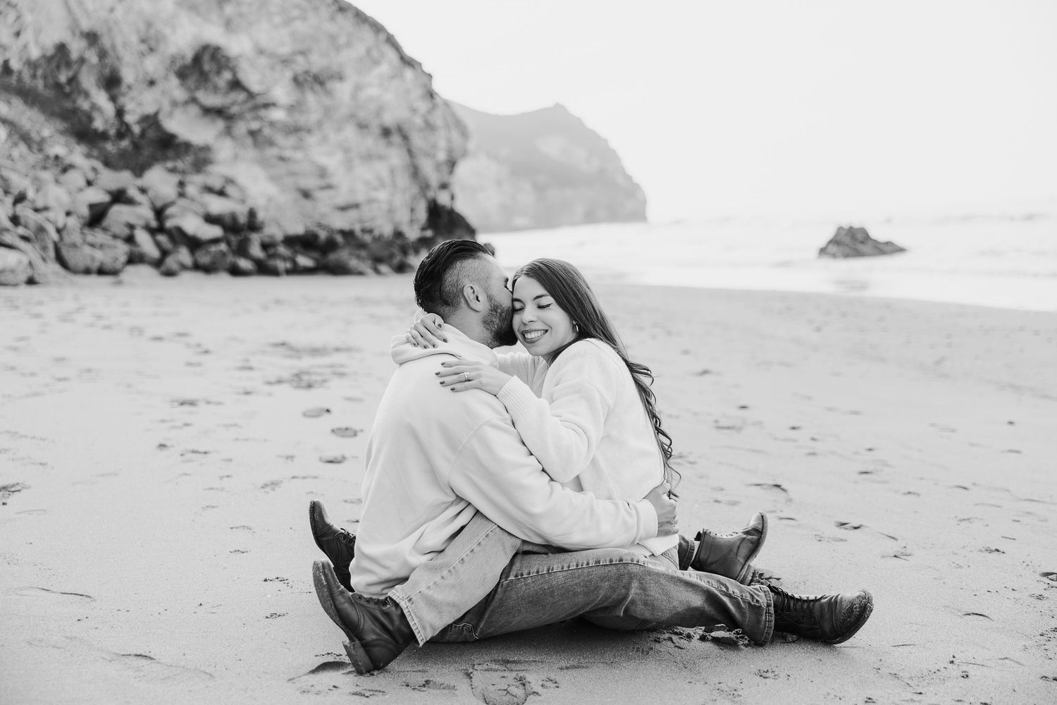 Black and white photo of couple relaxing on sandy beach near dramatic coastal rocks.