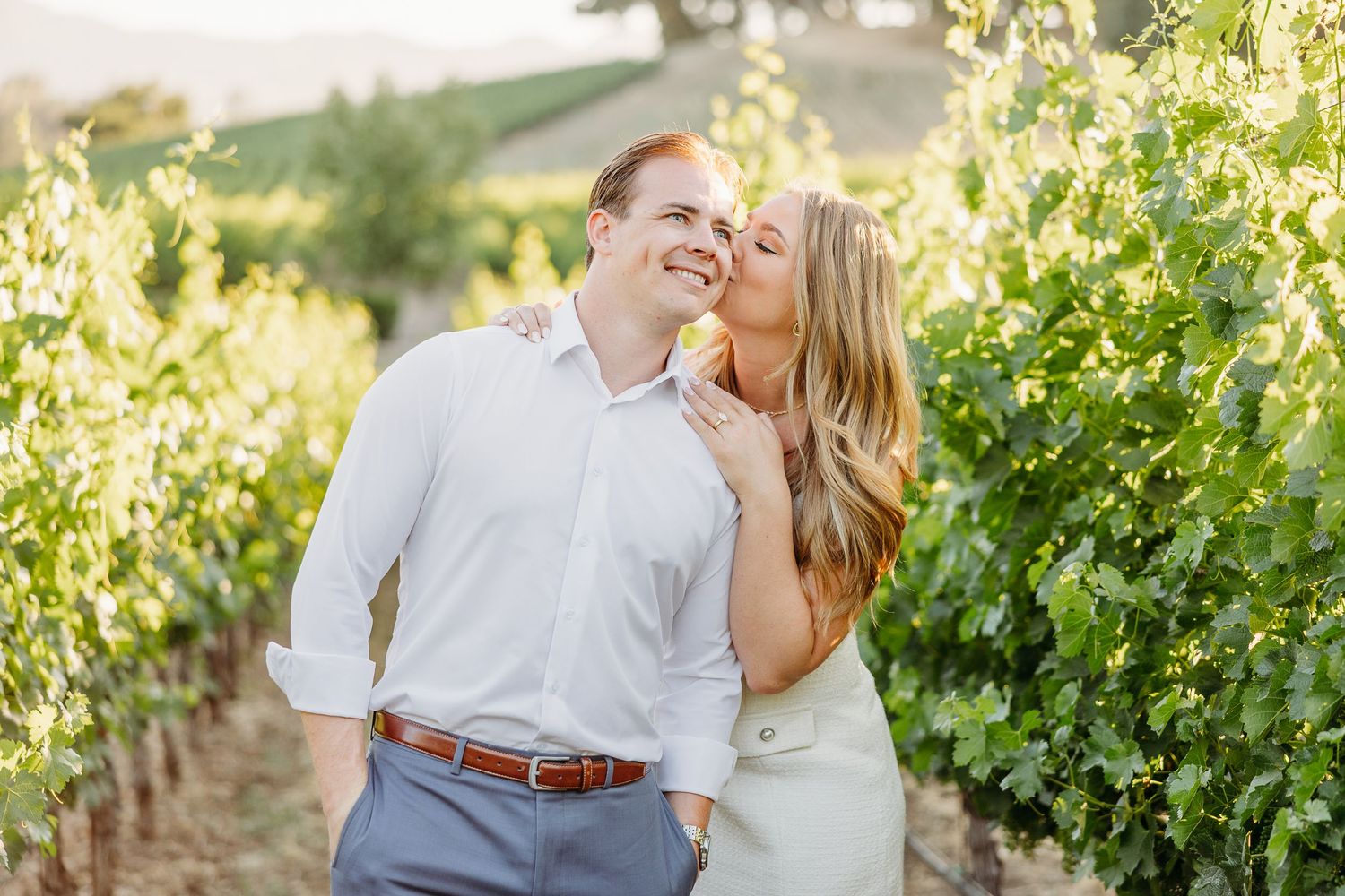 A couple shares a romantic moment among lush green grapevines in a sunlit vineyard.