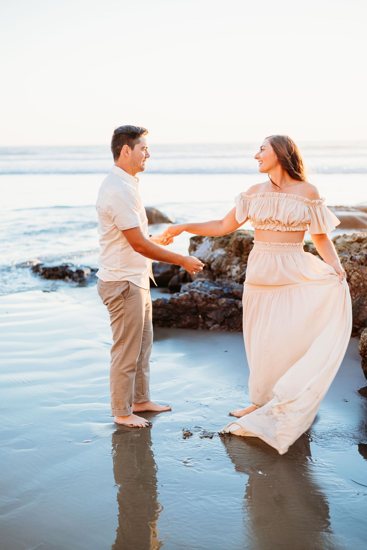 Couple dancing barefoot in shallow ocean water during golden hour near coastal rocks.