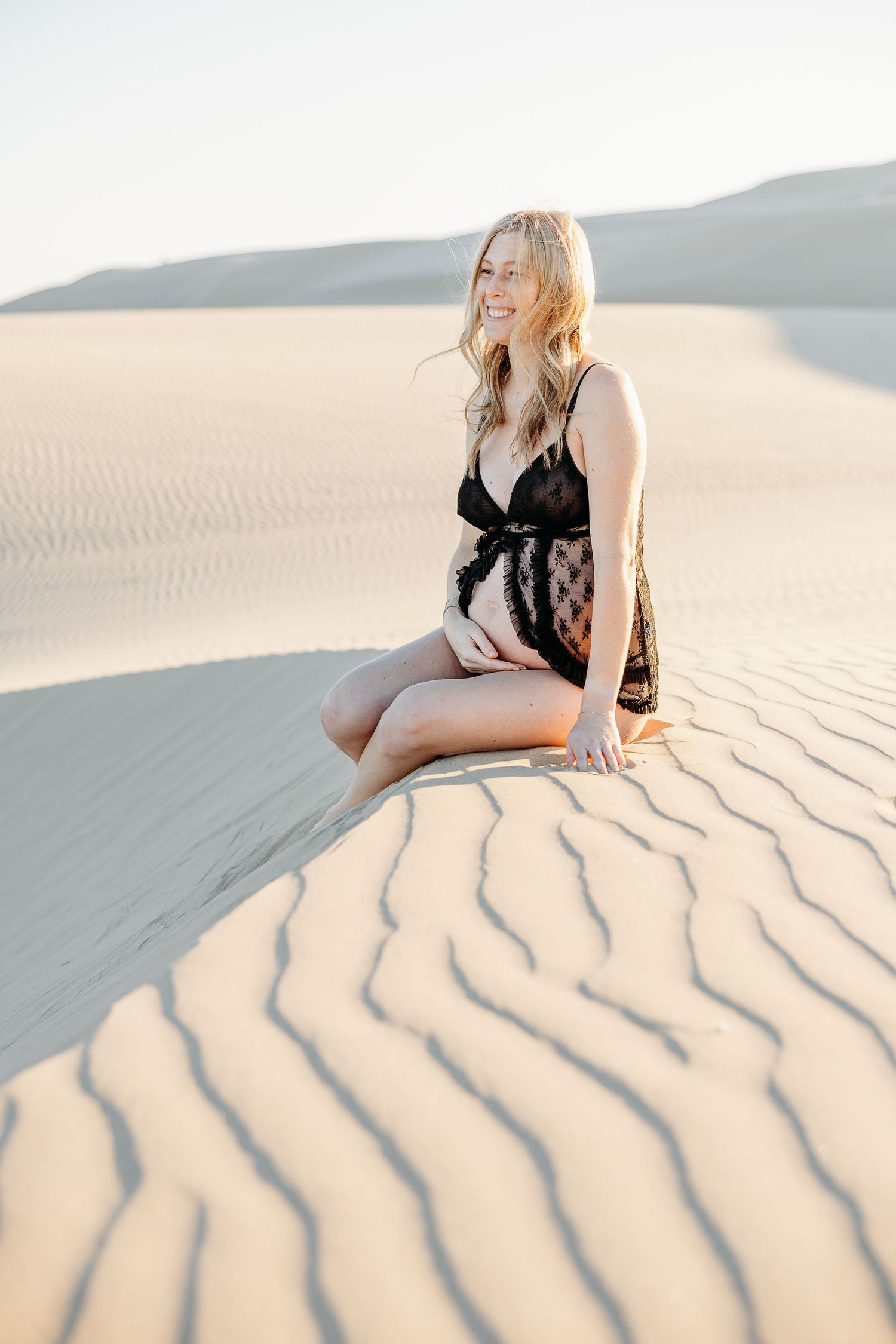 Person in black lace loungewear sits on textured sand dunes during golden hour.