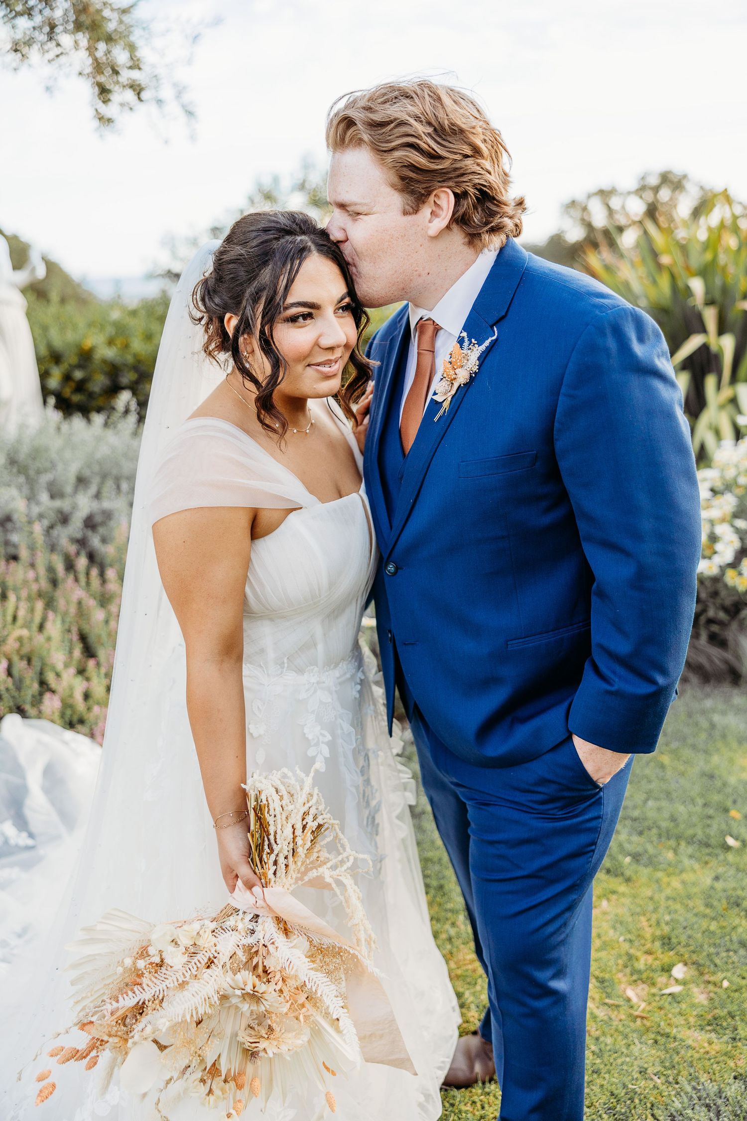 Wedding portrait with bride in white dress and groom in blue suit holding dried bouquet.