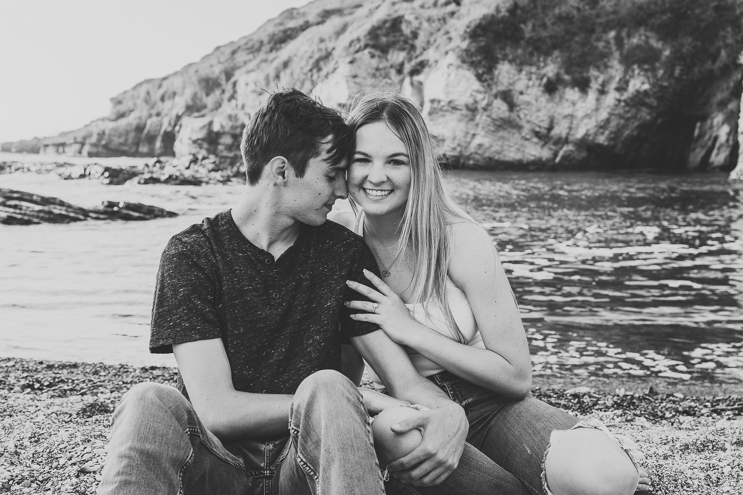 A black and white photo of a couple laughing together on a beach with waves behind them.