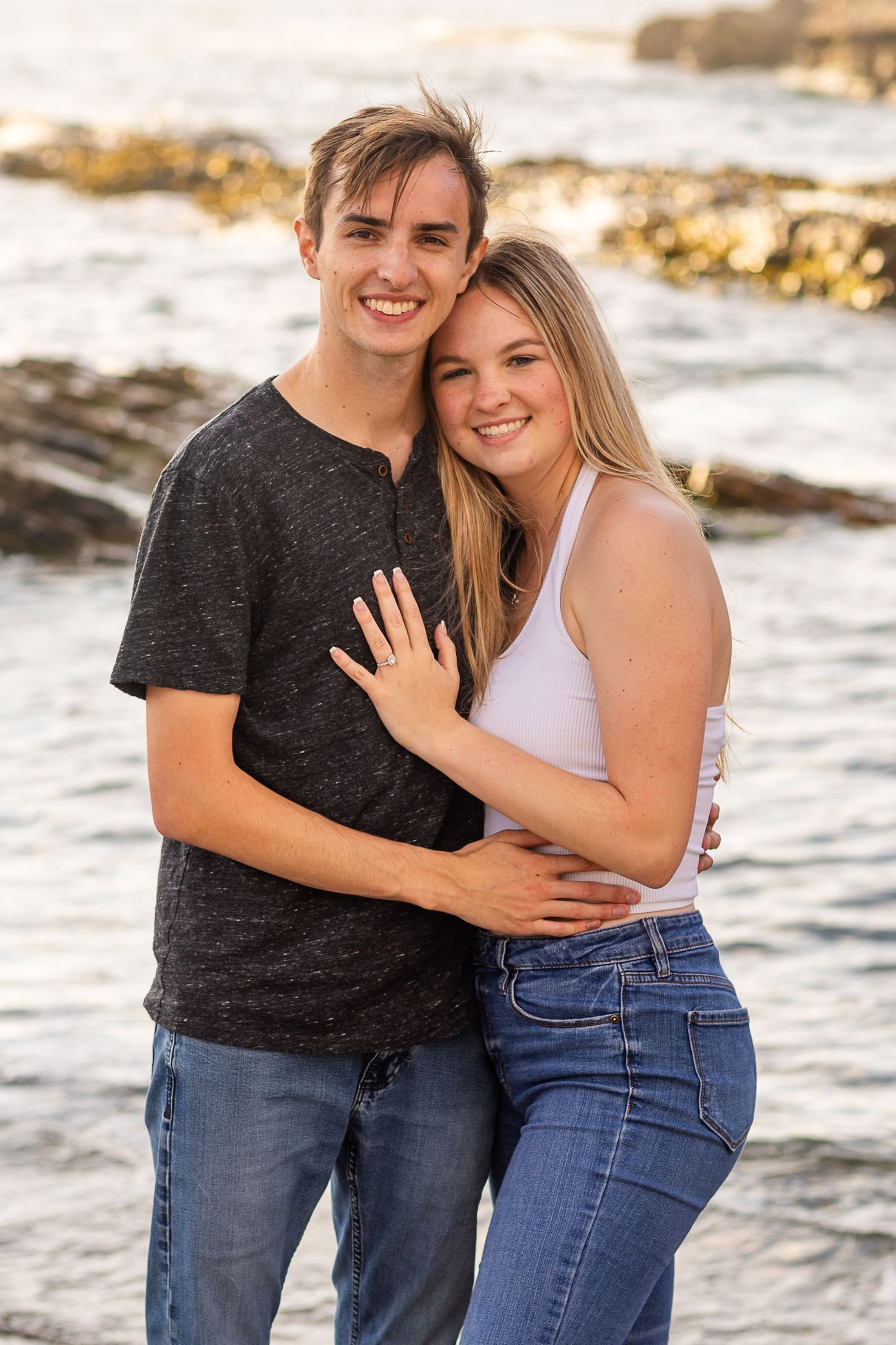 A couple poses together on rocky shoreline with ocean waves in the background.