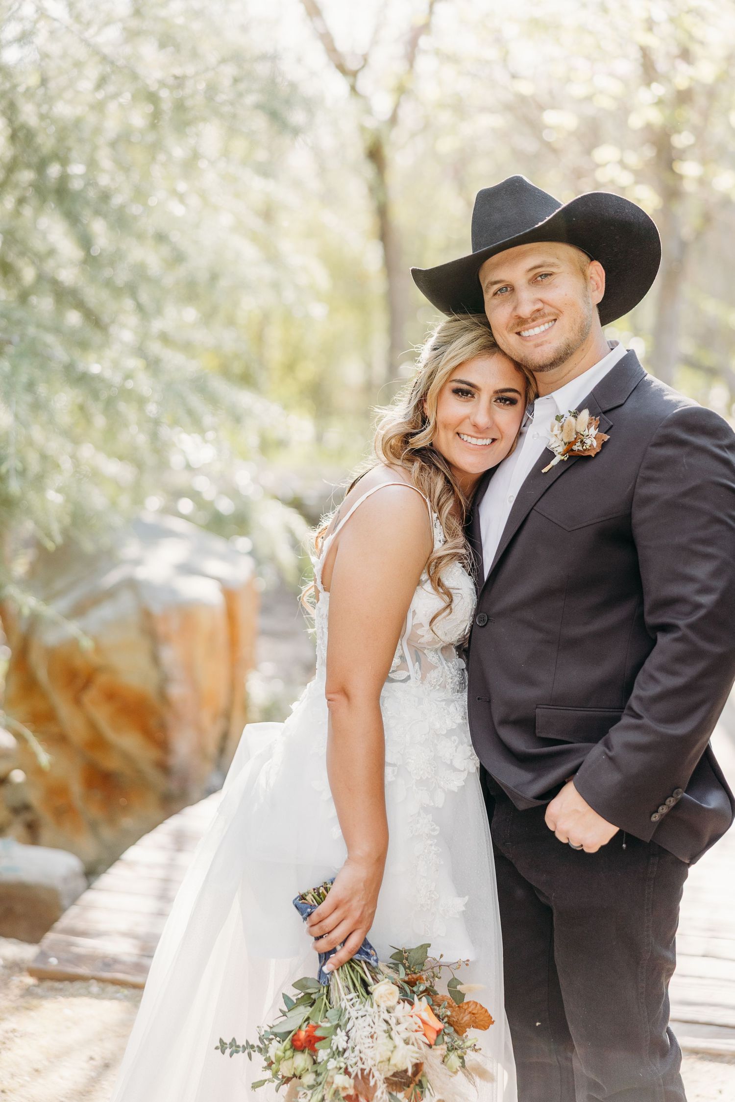 A western-themed wedding portrait of a couple in cowboy hat and white dress against natural scenery.