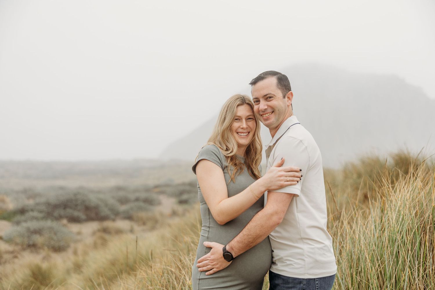 An expecting couple laughs together while embracing on sandy dunes with beach grass in the background.