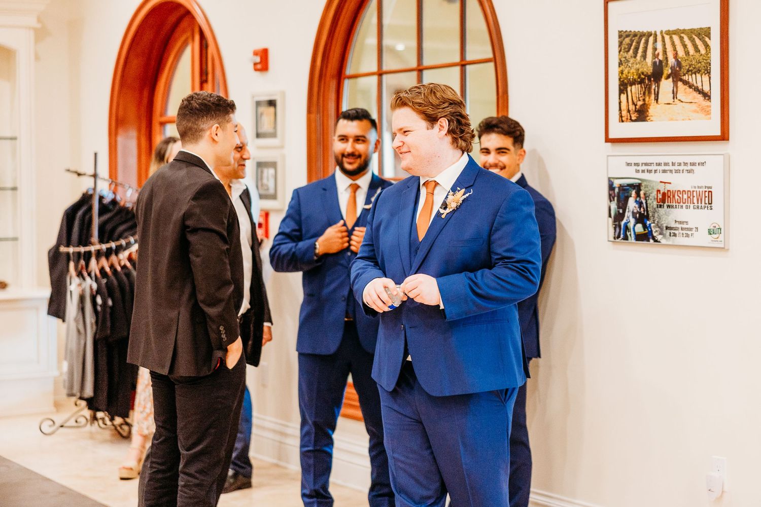 Wedding party members in suits chat in a bright hallway with arched doorways.
