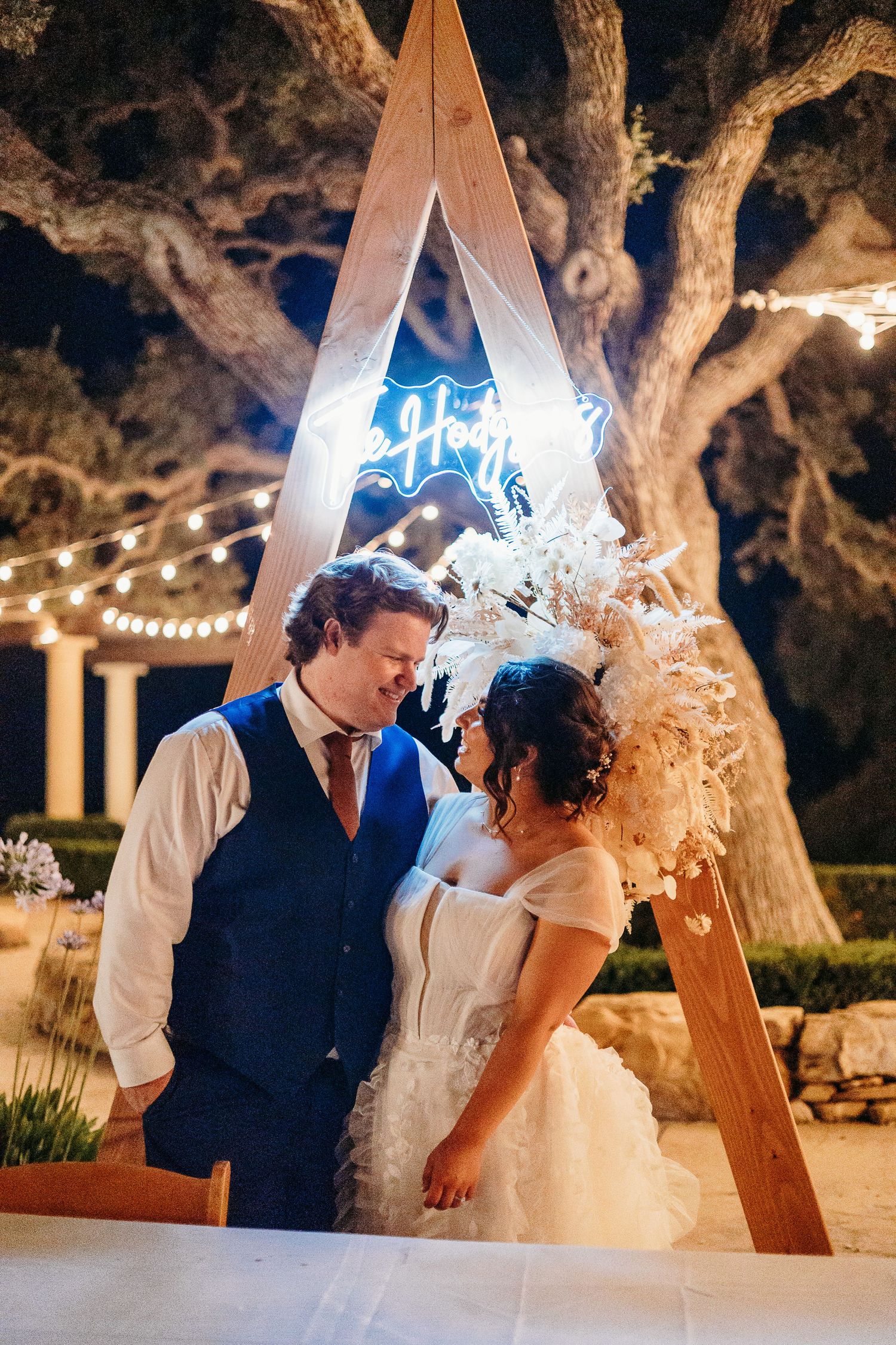 Intimate moment between couple under illuminated wooden arch at nighttime wedding.