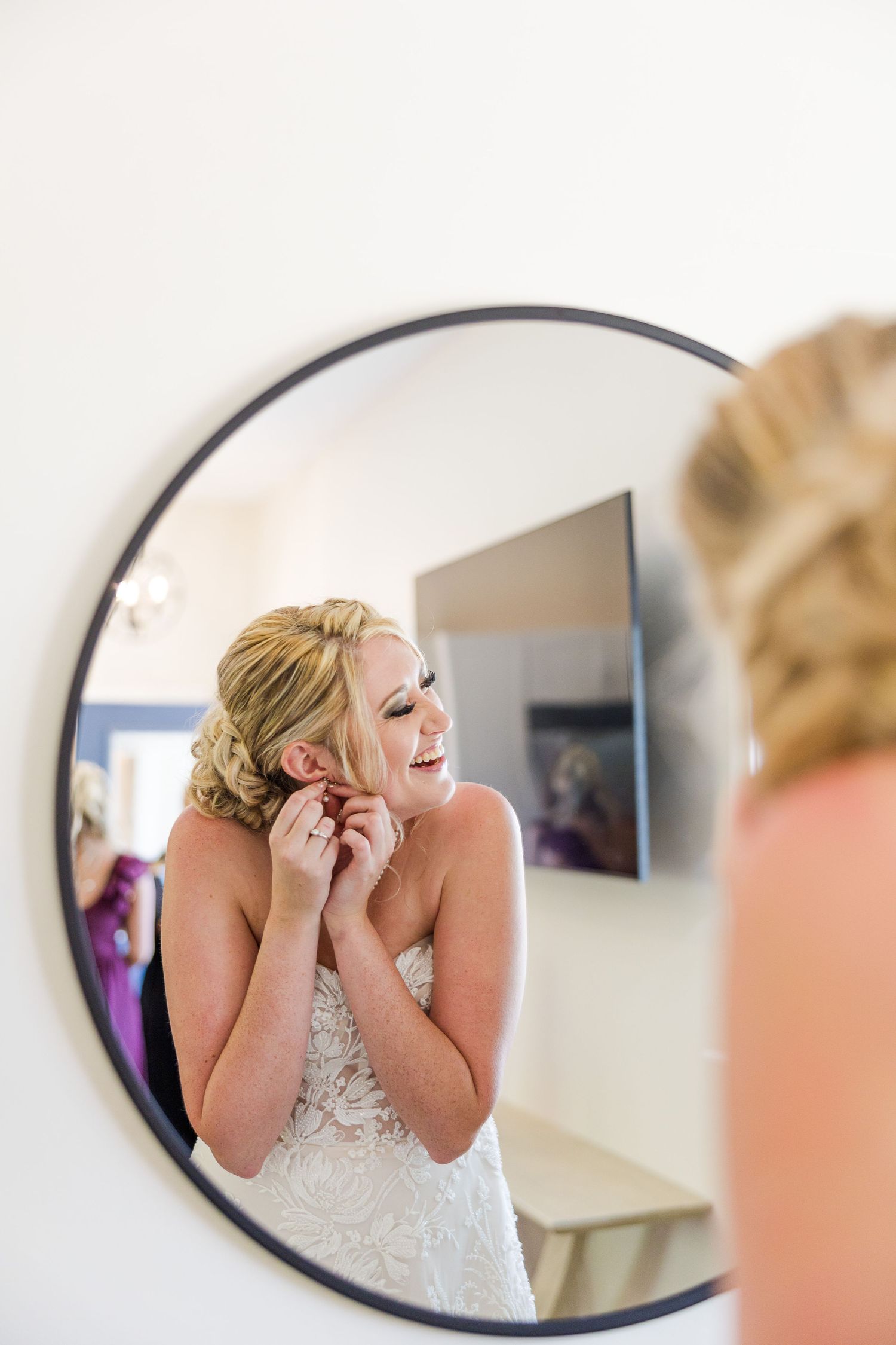Joyful reflection captured in round mirror shows bride wearing lace gown and elegant updo.