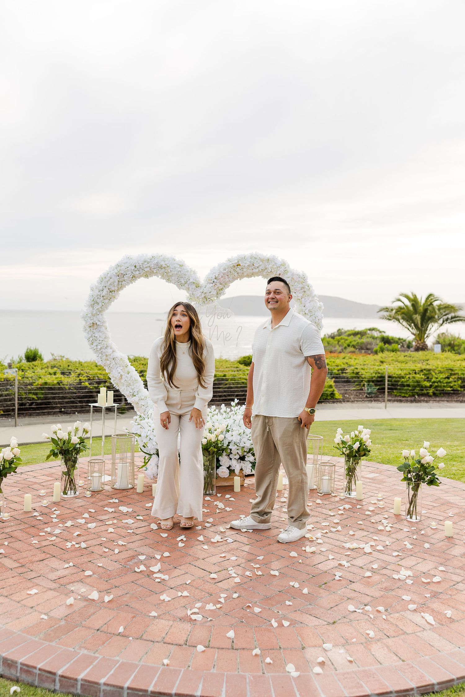 A wedding ceremony setup on a brick patio with white rose petals scattered in a heart shape and palm trees in background.