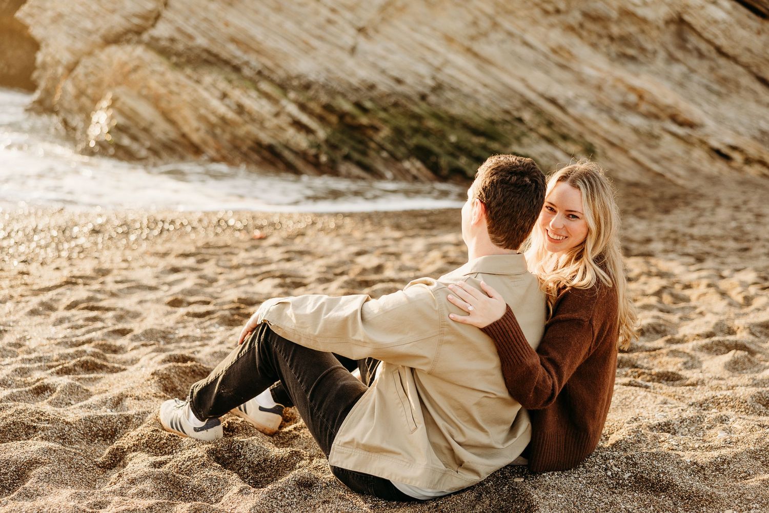Couple relaxing and laying on sandy beach near coastal rocks at golden hour.