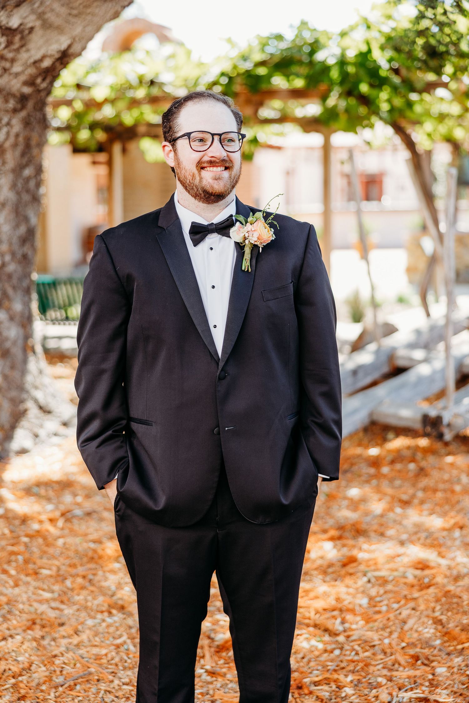 A groom in classic black tuxedo with bow tie and boutonniere poses outdoors.