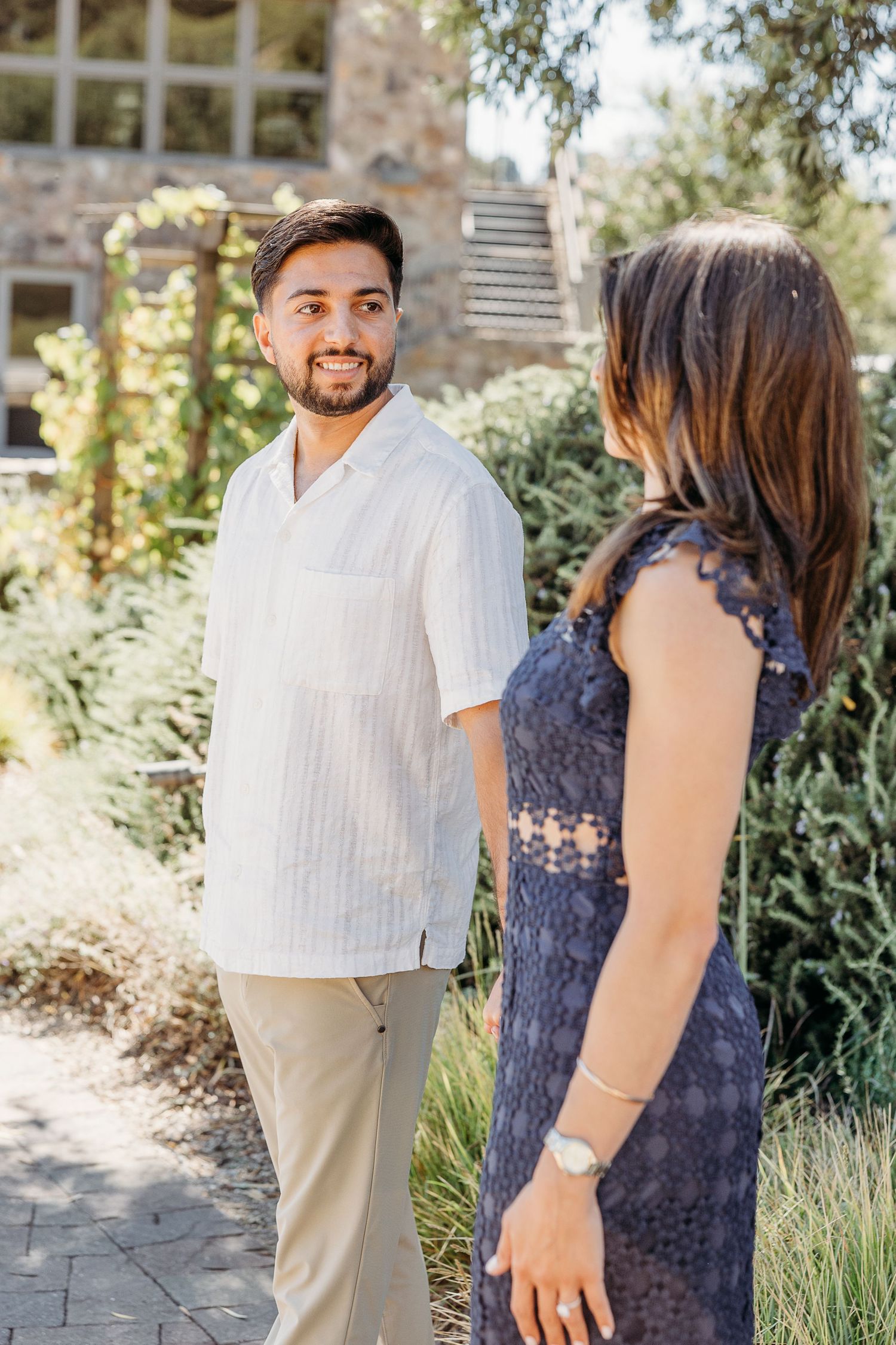 Couple walking along a garden path beside a stone building on a sunny day.