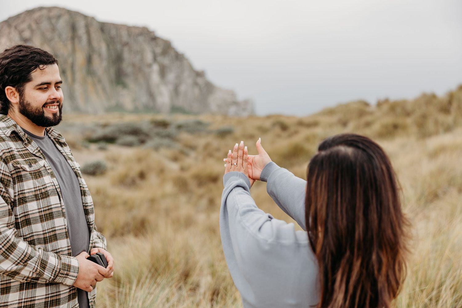Two people share a joyful moment in dry grass field with dramatic cliffside mountain in the background.