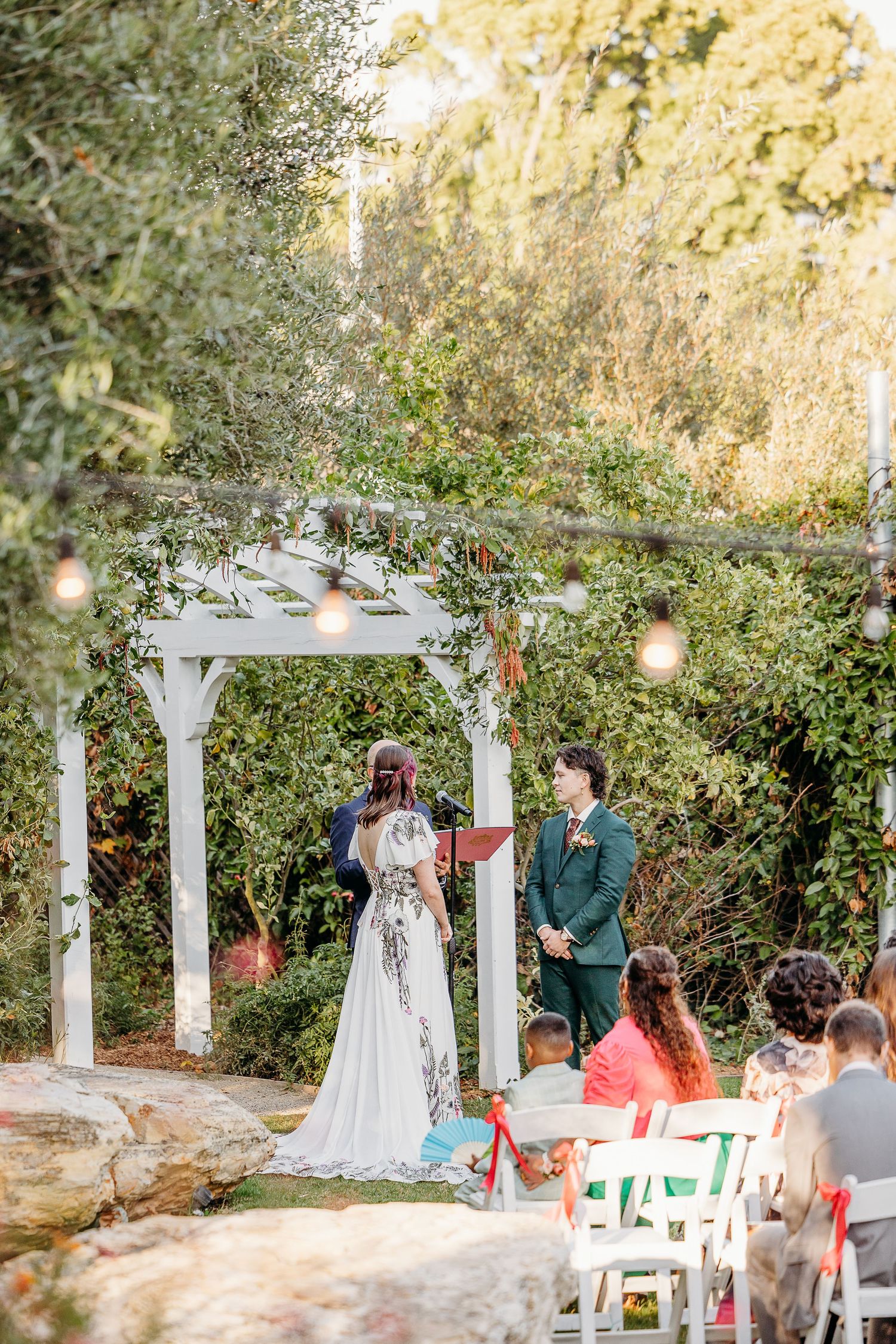 An outdoor wedding ceremony under a white pergola decorated with greenery and flowers as guests sit in white chairs.