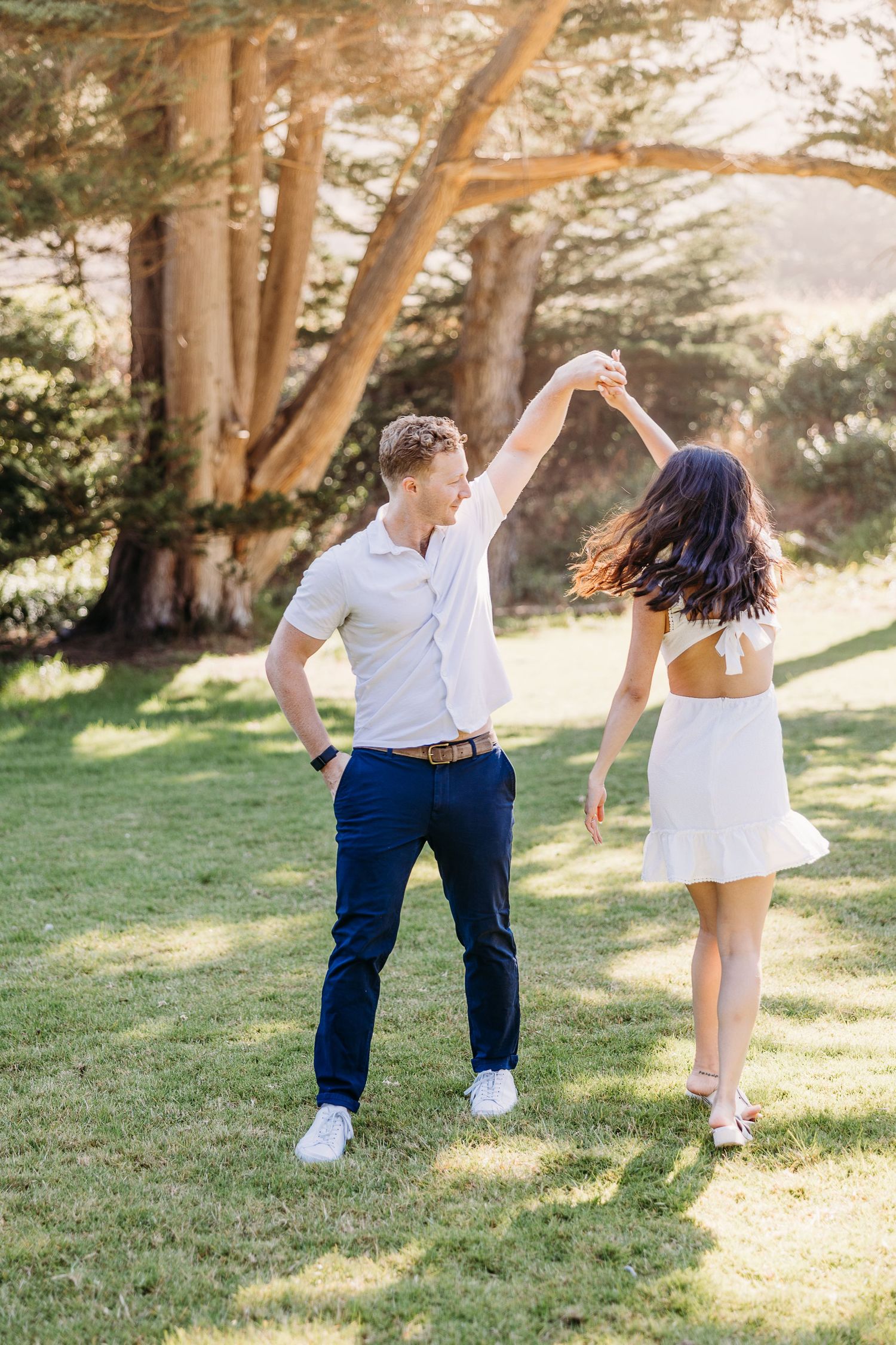 Couple dancing playfully on grass with tall trees in background.