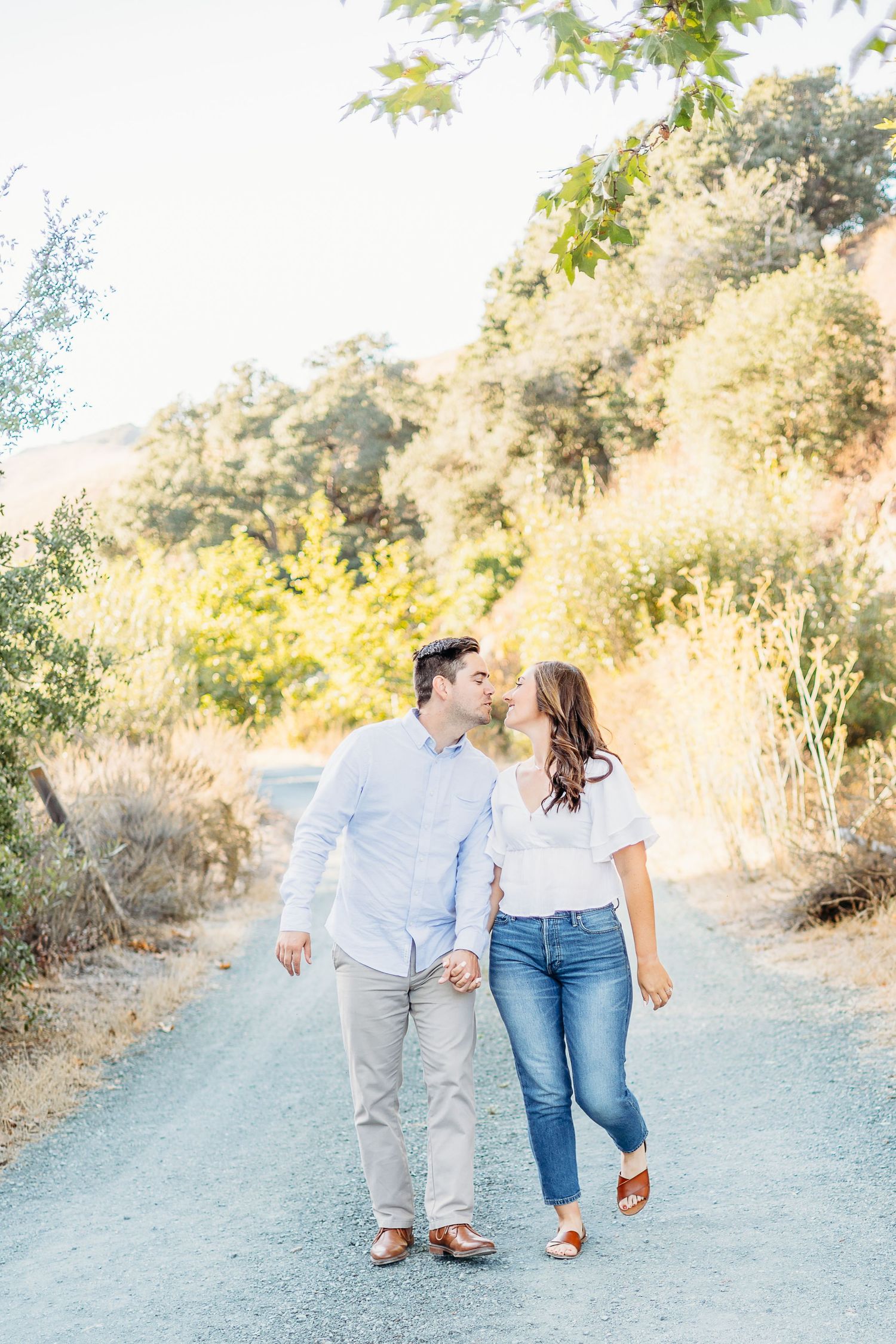 A couple walks hand in hand along a dusty path surrounded by golden sunlit trees.