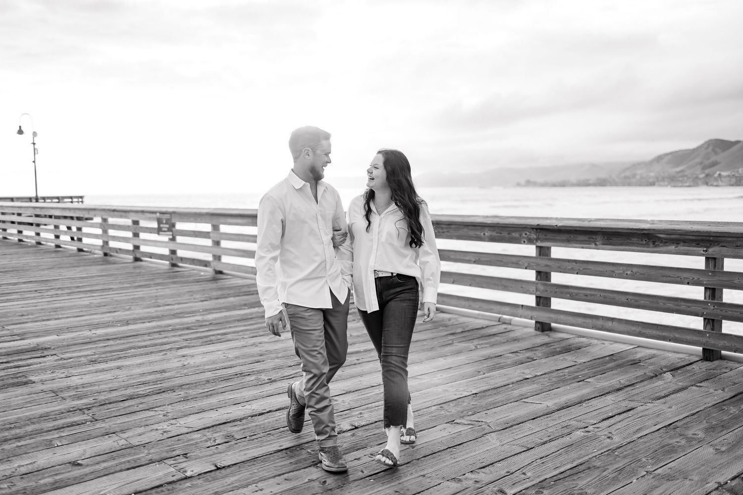 A black and white photo of a couple walking together on a wooden pier by the ocean.
