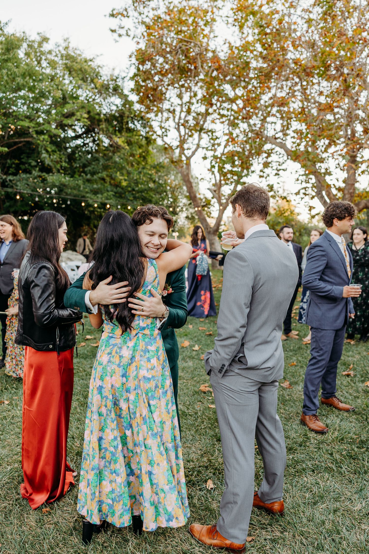 Wedding guests in colorful dresses and suits embracing during outdoor reception.