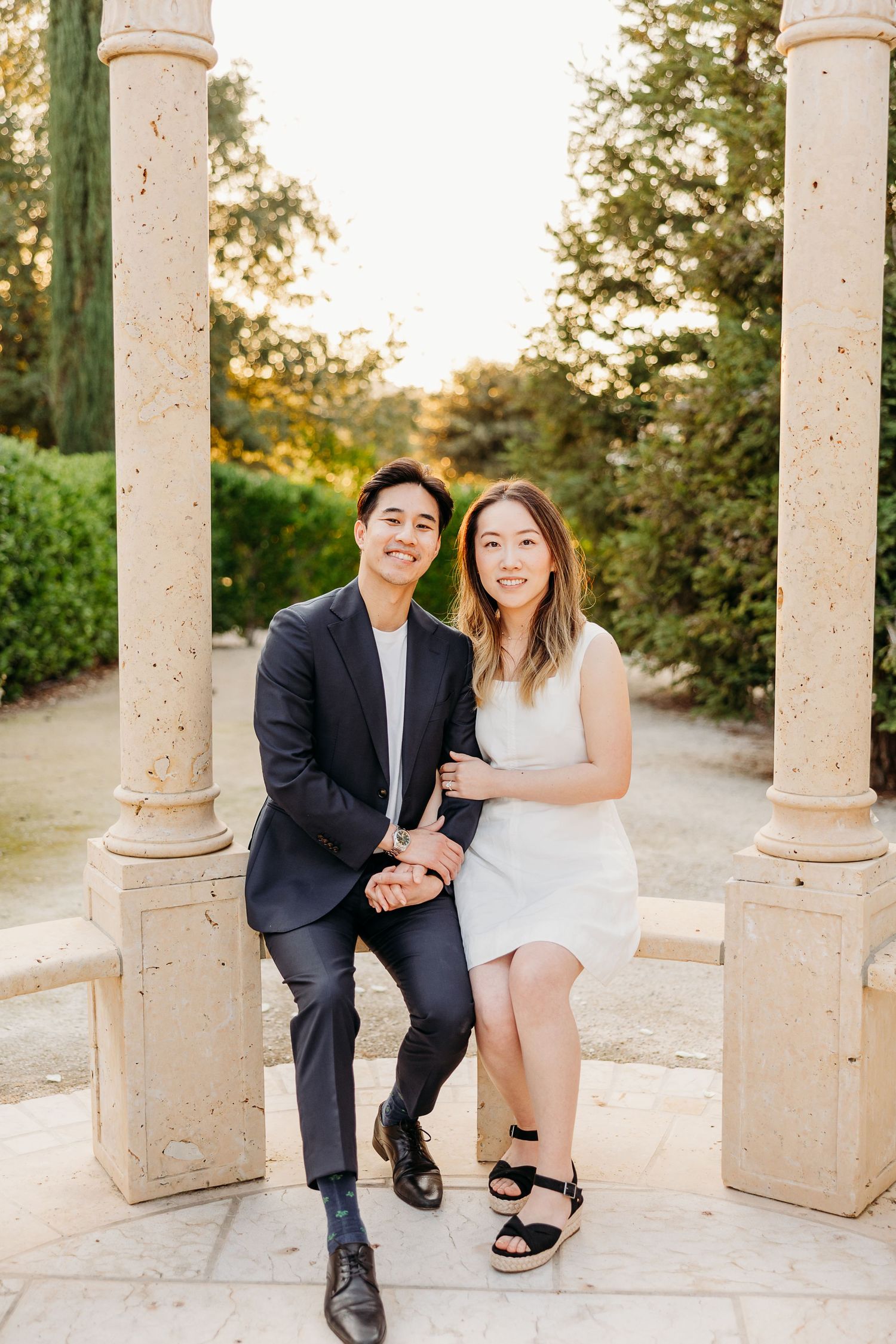 A couple sits between stone pillars in a garden setting during golden hour.
