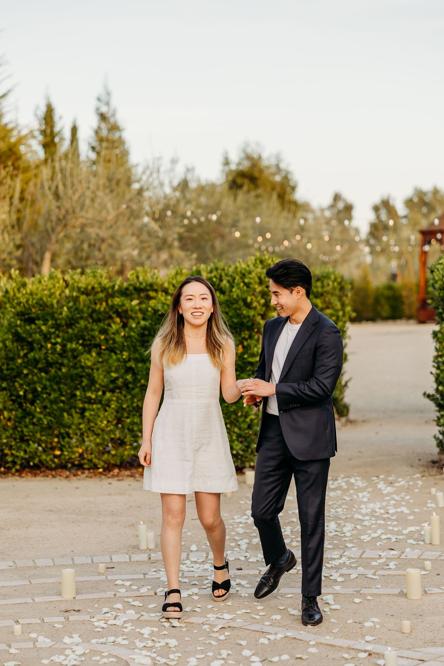 A couple walks together laughing on a stone path surrounded by greenery and white floral decorations in the garden at Allegretto.