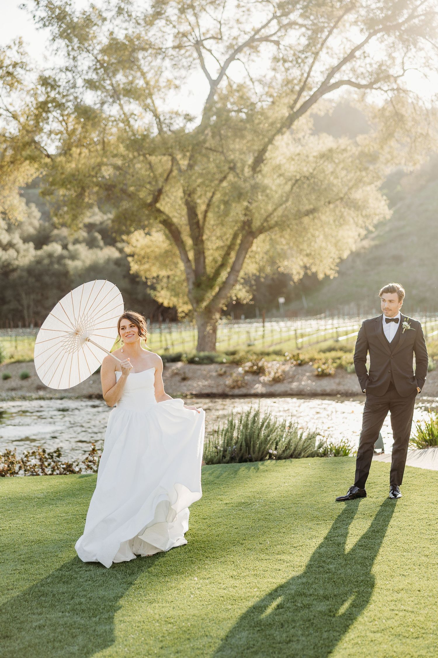 A couple in wedding attire poses playfully with a white parasol on a sunlit lawn beside a pond.