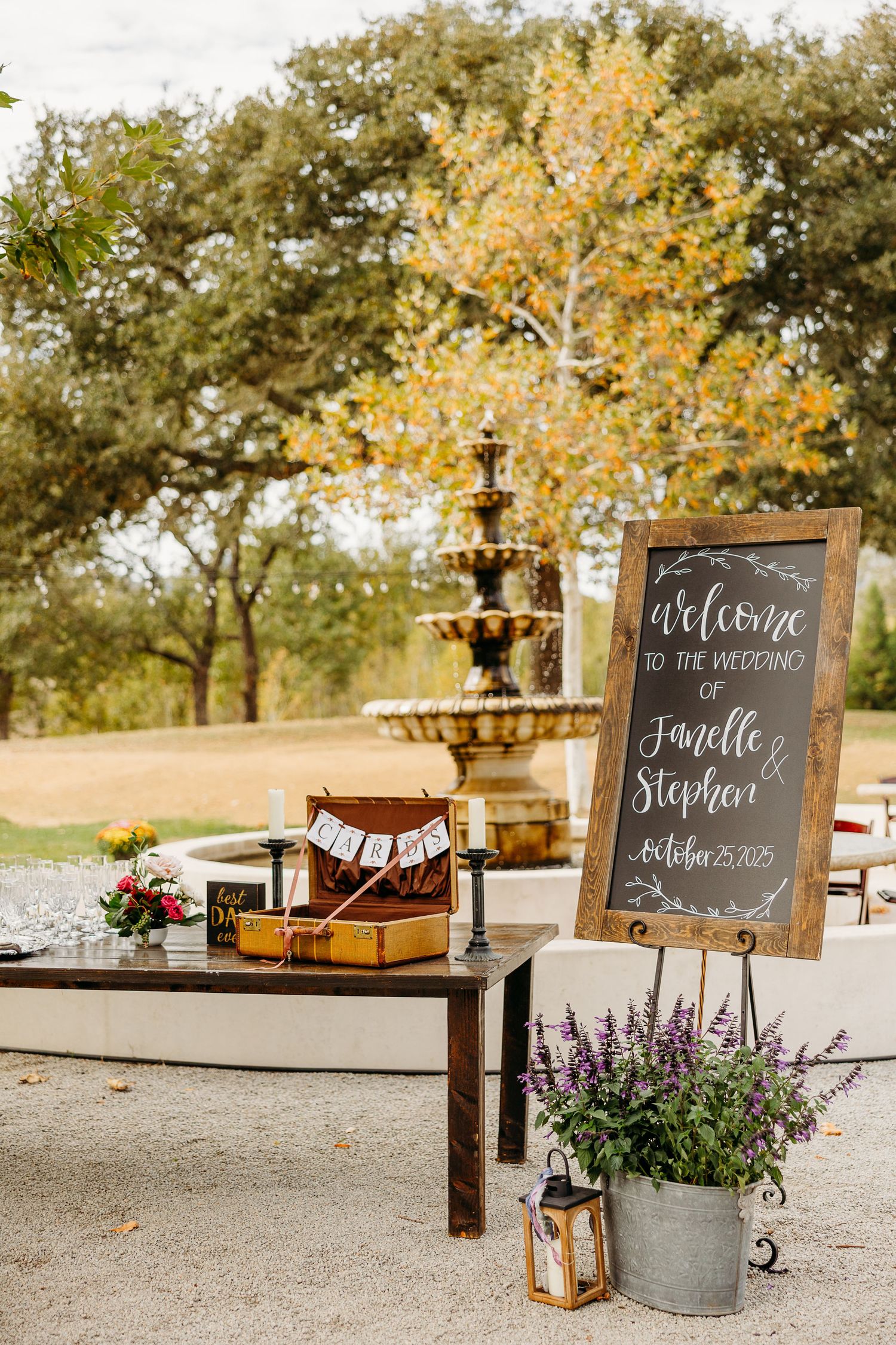 Rustic wedding welcome display with chalkboard sign, vintage suitcase, and tiered fountain against autumn trees.