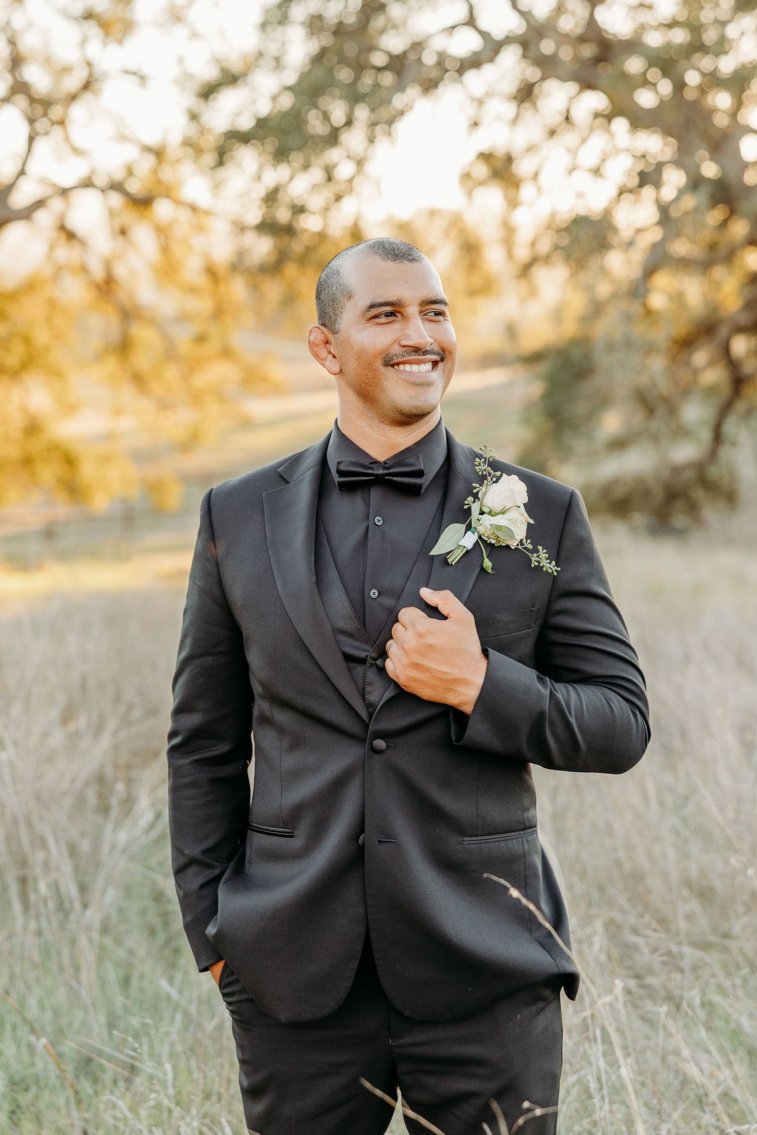 Groom in black tuxedo smiling while adjusting boutonniere outdoors.